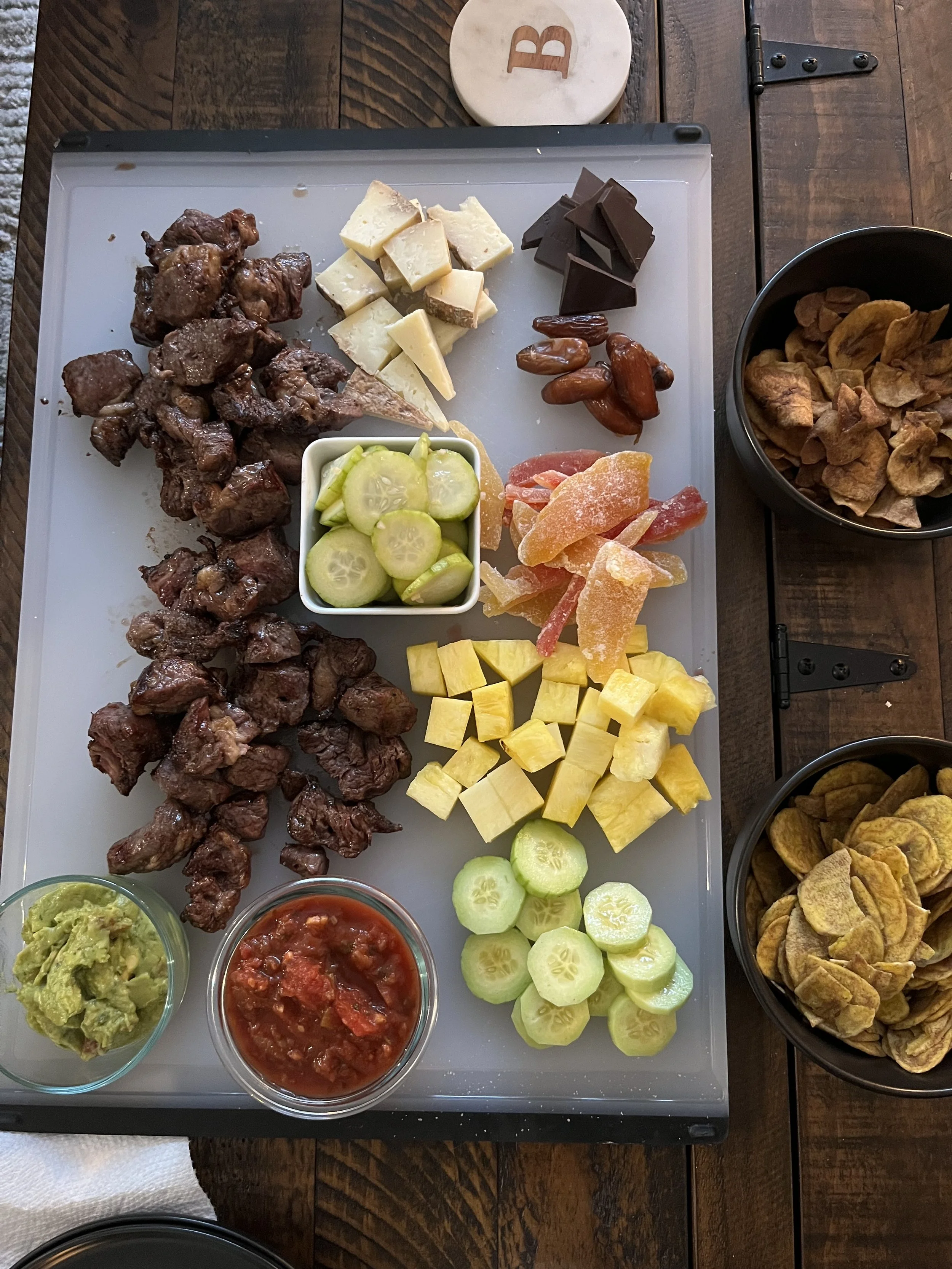A tray of assorted foods including grilled meat, various cheeses, pickles, dried dates, candied ginger, sliced fruit, guacamole, and salsa, with bowls of chips, guacamole, and salsa.