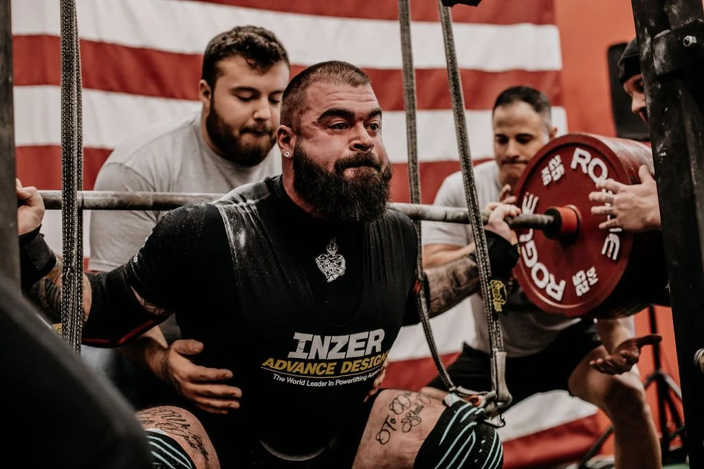 A man with a beard and tattoos is squatting and holding a barbell with red weights during a powerlifting competition. Two spotters assist him while a flag with red and white stripes hangs in the background.