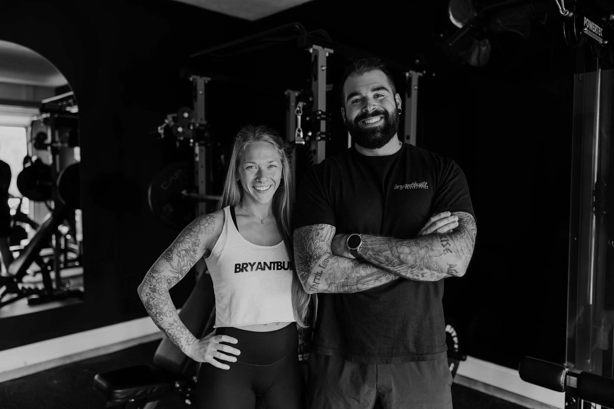 Two smiling individuals, a woman and a man, standing side by side in a gym, with gym equipment in the background. The woman has long hair, tattoos on her arms, and is wearing a sleeveless top with "BRYANTBUILD" written on it. The man has a beard, tattoos on his arms, and is wearing a black T-shirt with the same logo.