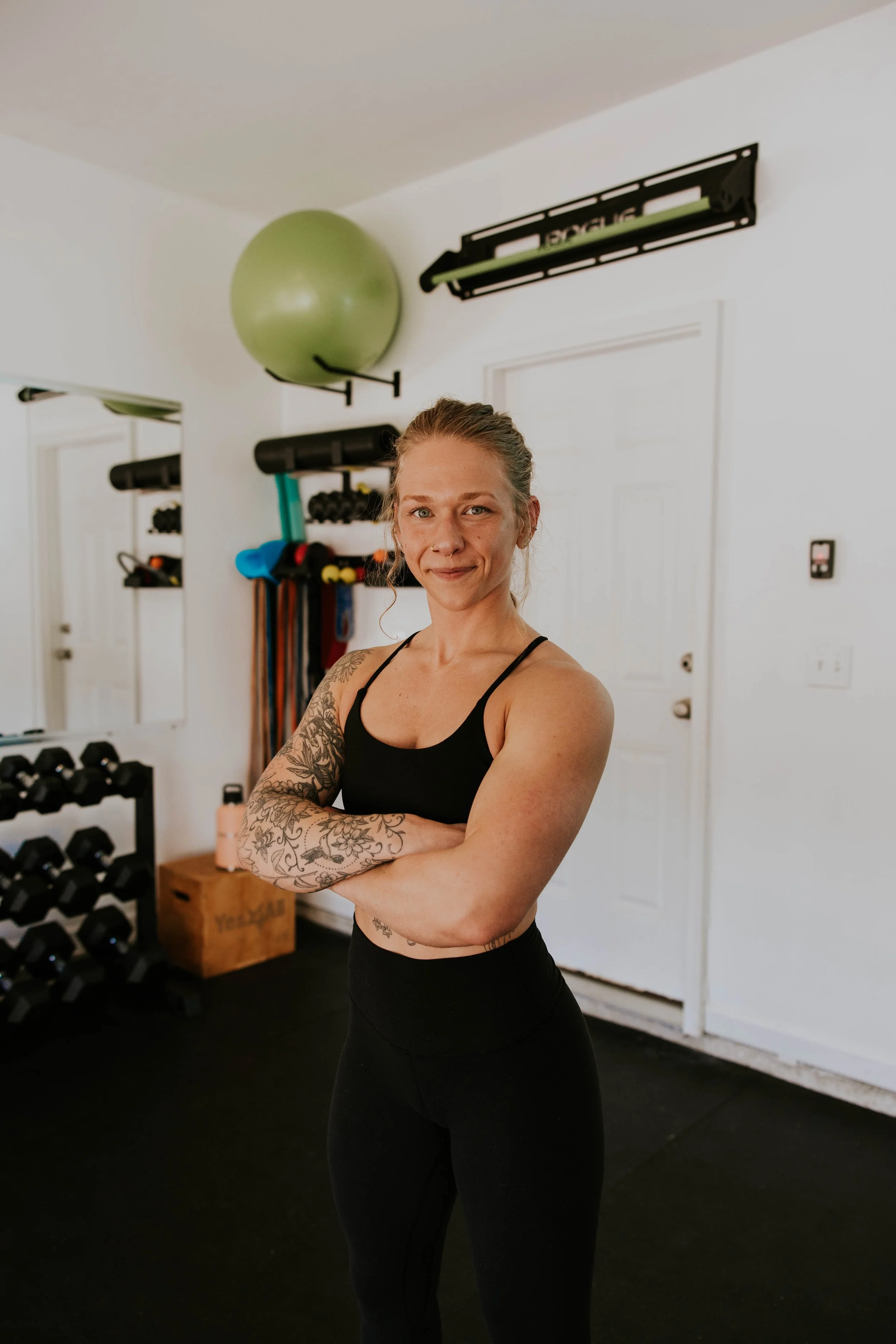 A woman with tattoos on her arm and a nose ring taking a selfie in a home gym. She is wearing black athletic wear and standing with her arms crossed. There are black dumbbells, a wooden box, a green exercise ball, and wall-mounted workout equipment in the background.