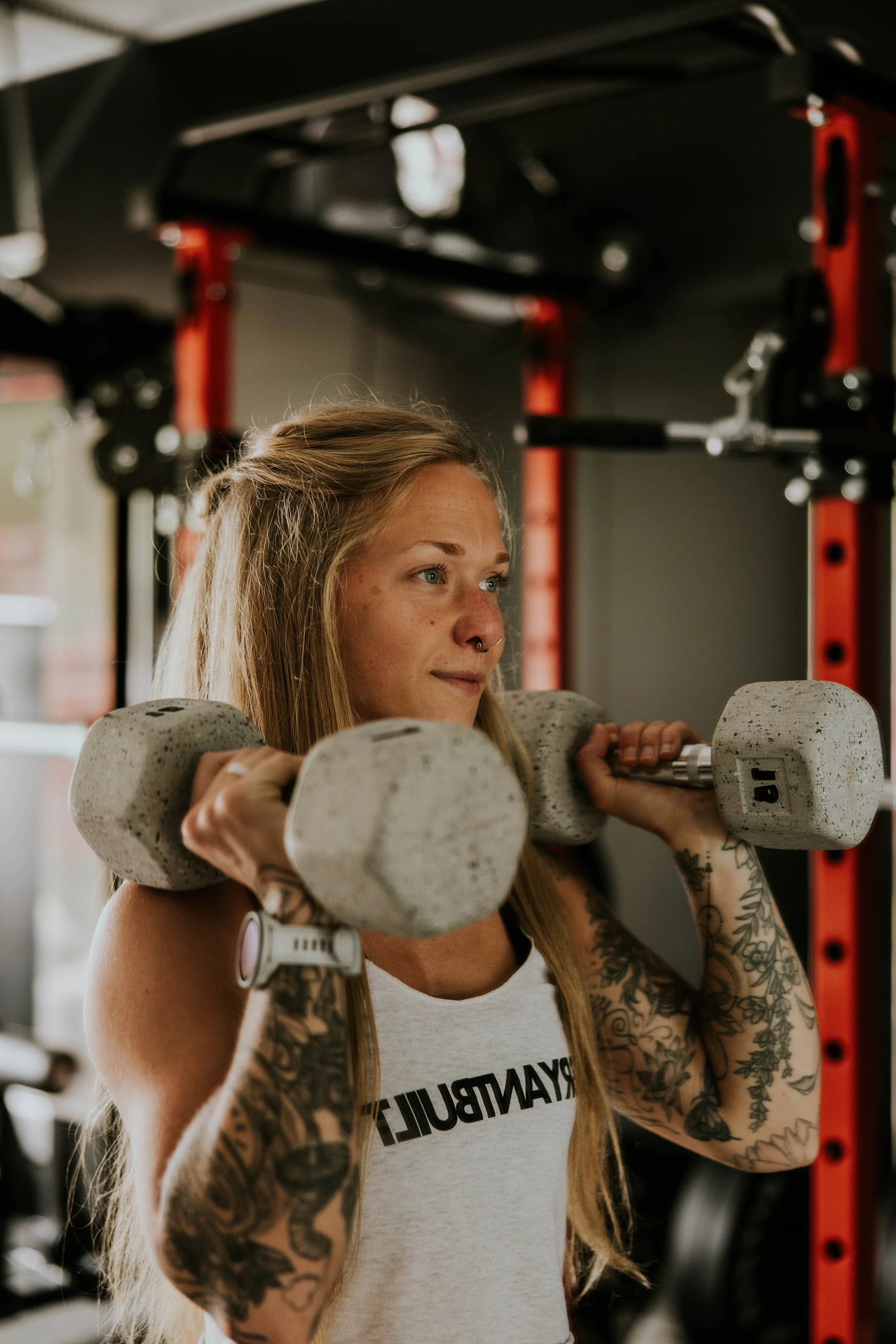 A young woman with long blonde hair, tattoos on her arms, and a septum piercing is lifting a dumbbell during a workout in a gym.