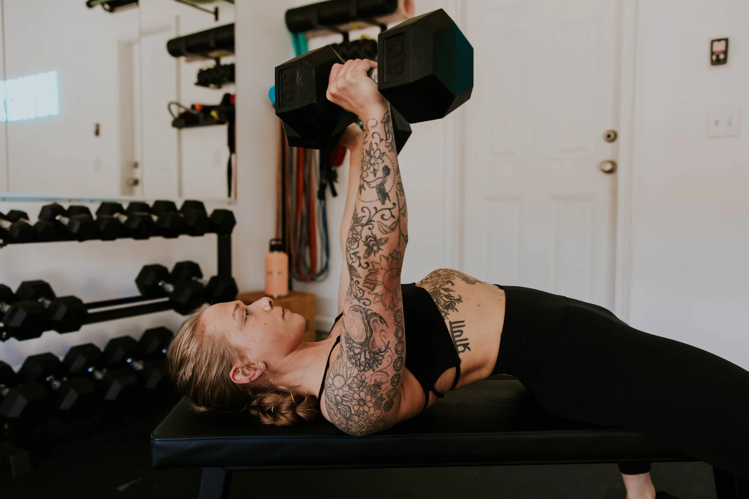 A woman with tattoos on her arms lying on a workout bench and lifting a dumbbell. She is in a gym with a rack of smaller dumbbells behind her and improvised workout equipment on the wall.