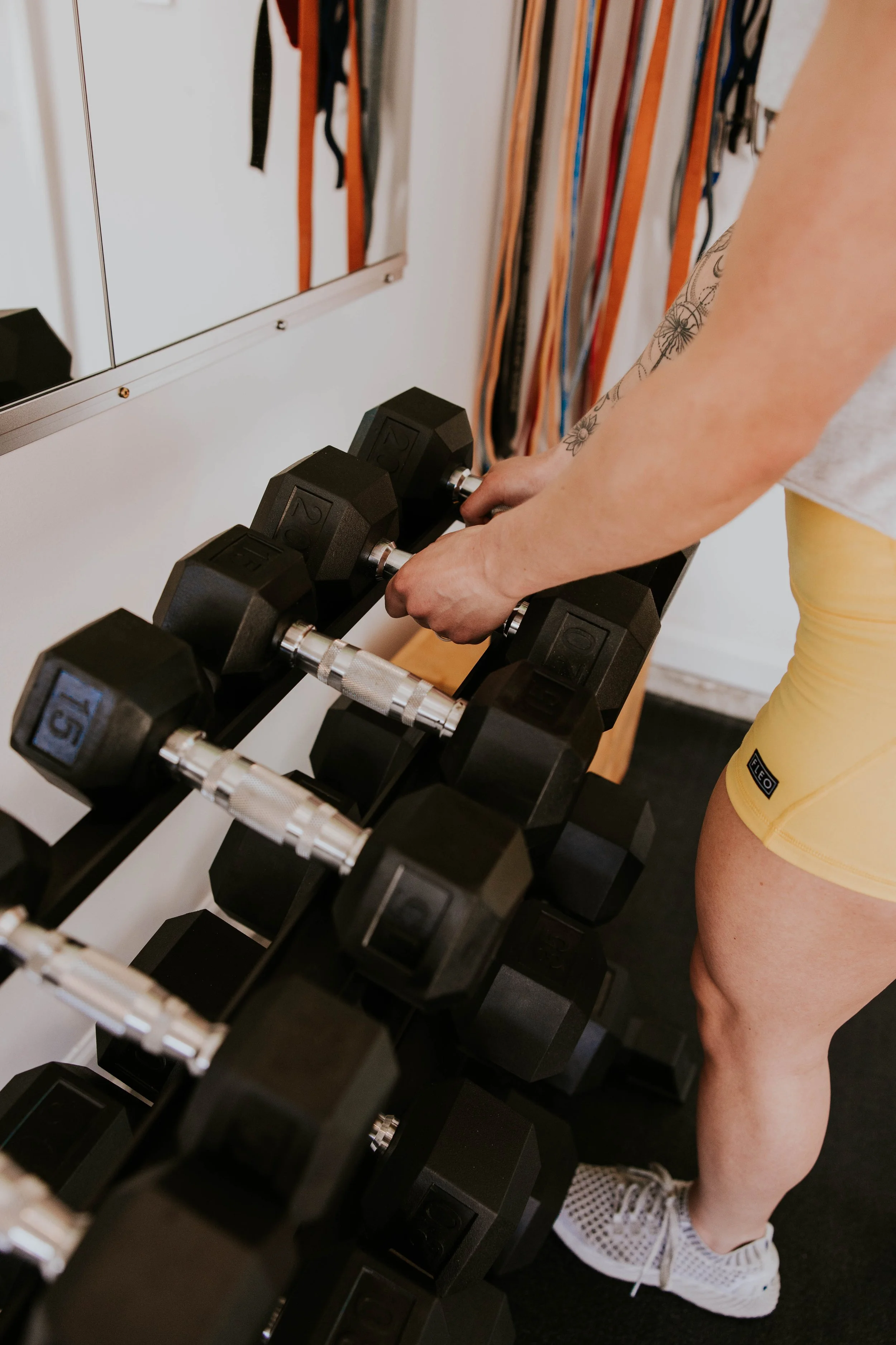 A person wearing yellow shorts, white sneakers, and a white shirt is holding dumbbells in a workout room. The person's tattooed arm is visible, and there are several black dumbbells on a rack.
