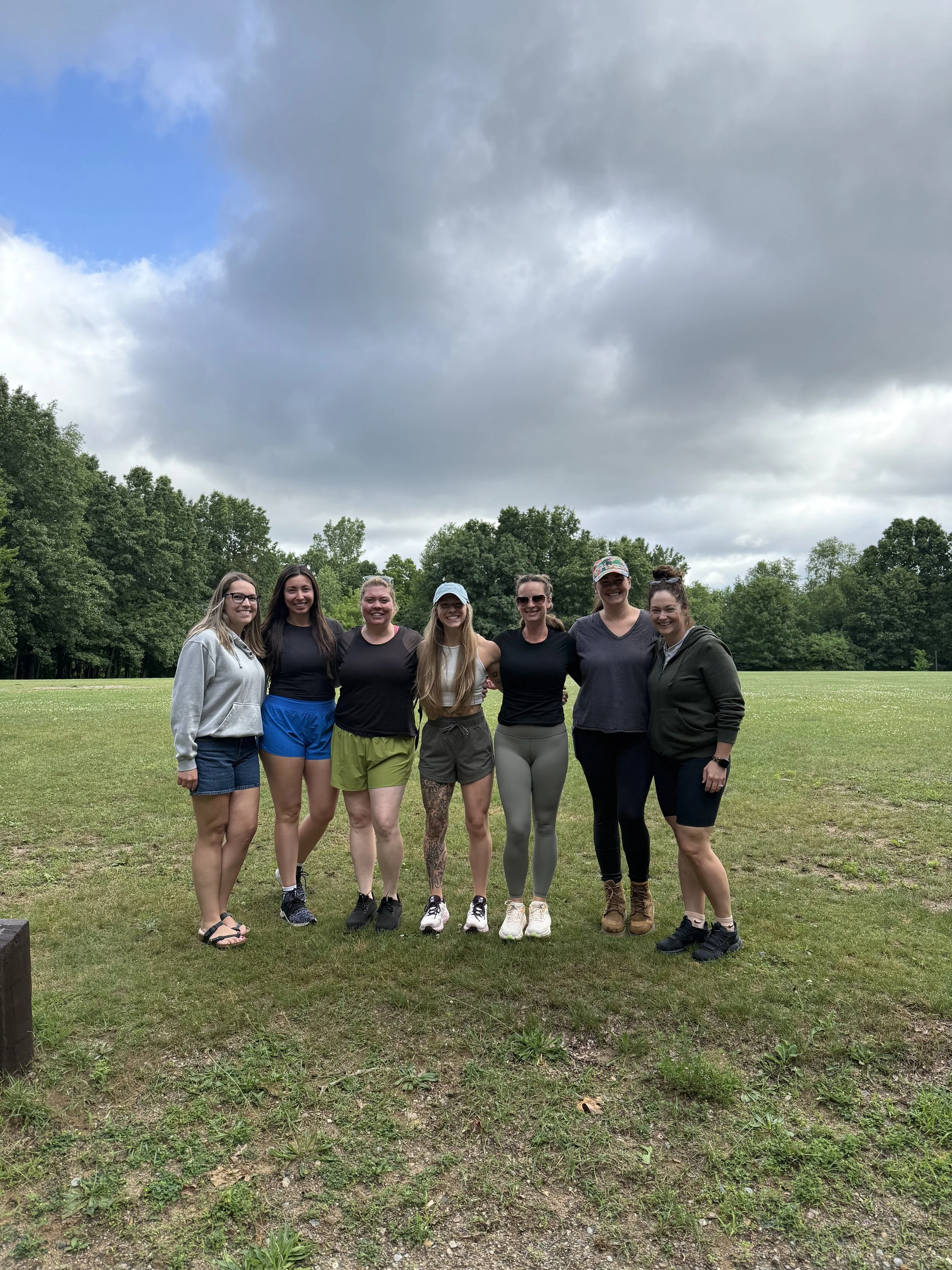 A group of seven young women standing outdoors on grass, with trees and cloudy sky in the background, smiling at the camera.