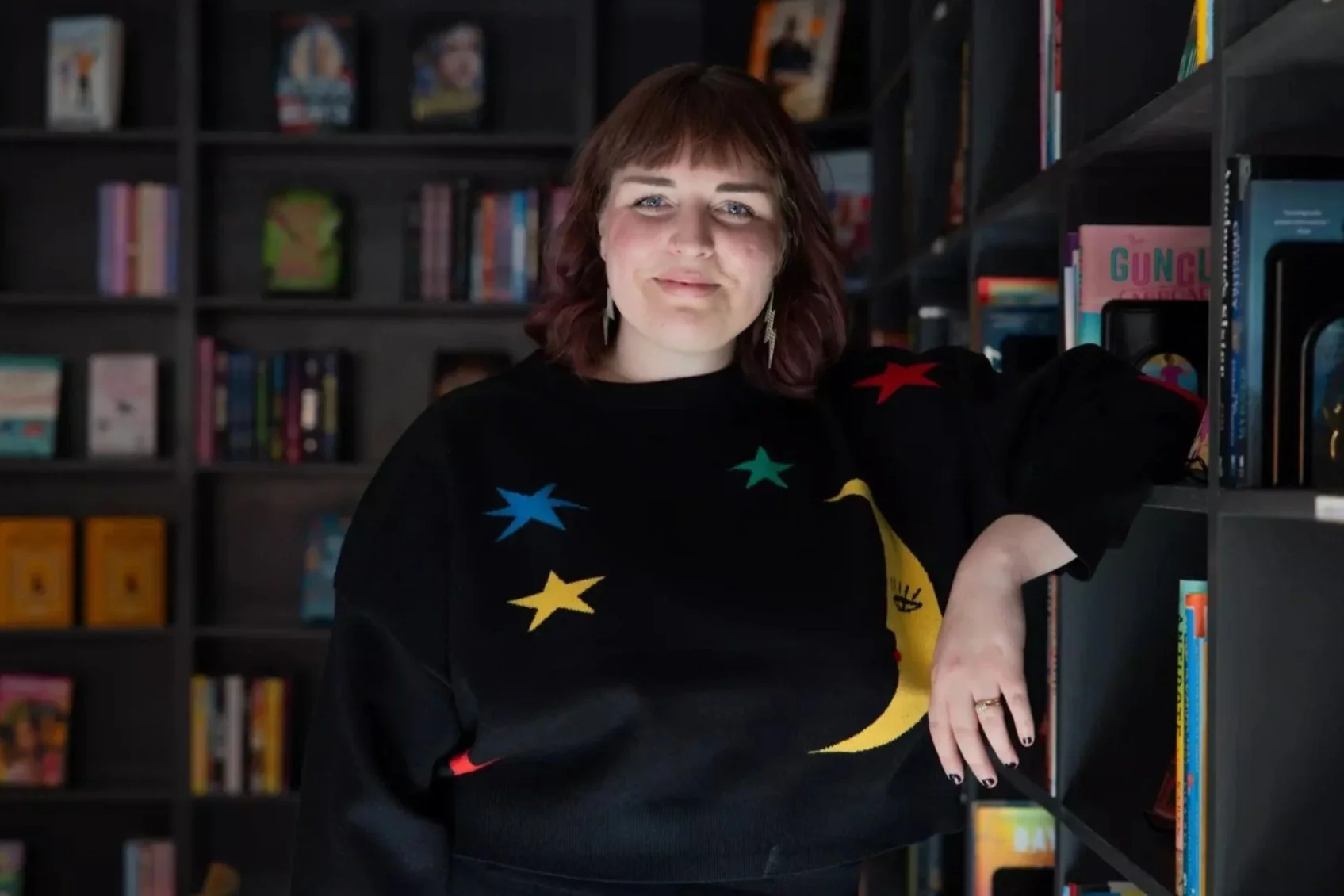 Caitlin Harrison stands and smiles in front of a stack of books, at Black Cat Fables in Monrovia, CA. She is wearing a La La Land black sweater set featuring colorful a moon and stars.