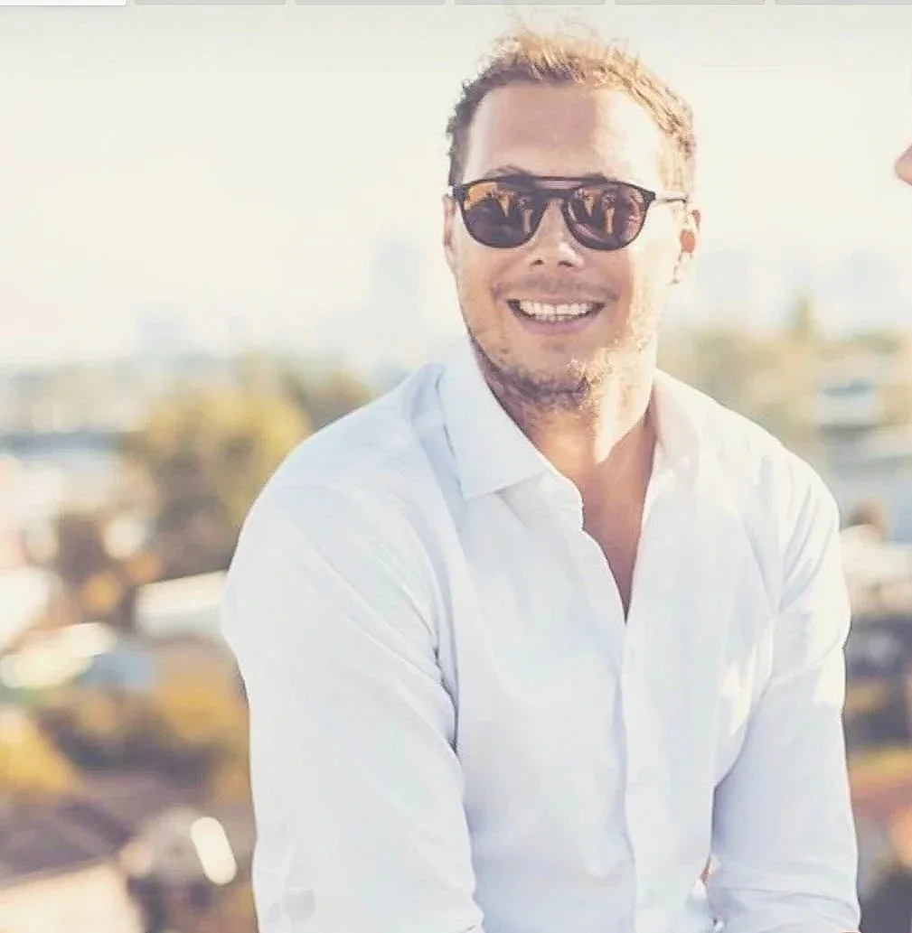 Ein lachender Mann mit Sonnenbrille und weißen Hemd, im Freien bei Sonnenlicht, mit einer urbanen Kulisse im Hintergrund.