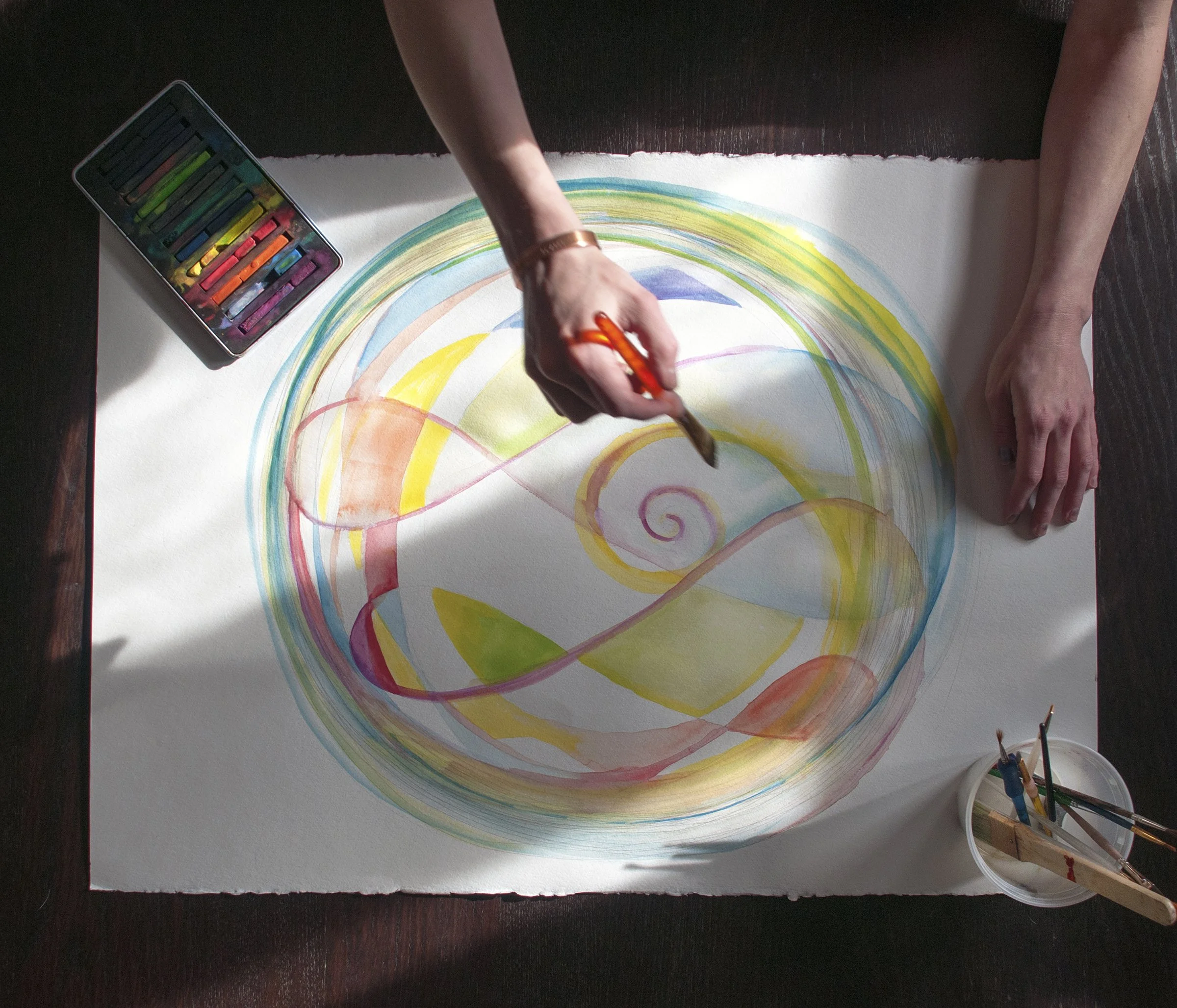 Person creating an abstract watercolor painting with swirling lines and colors on white paper. The person is using a paintbrush, and there is a set of watercolors and brushes nearby.
