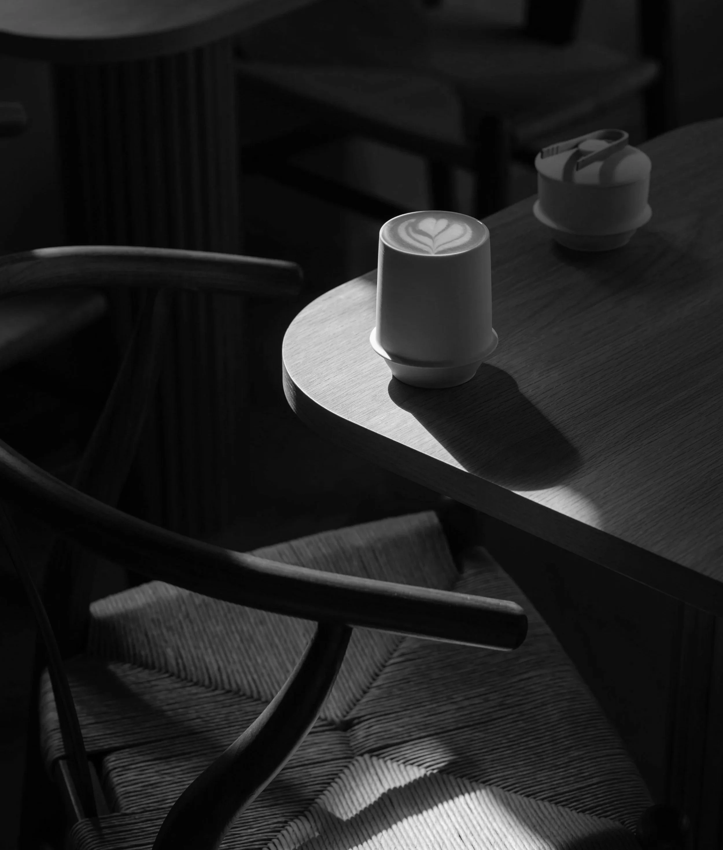 Close-up of a latte with latte art in a cup, on a wooden table. In the background, there is a small container with stir sticks or straws, and a wooden chair.  Speciality Coffee Bakery Near Me. London, Lake Como, Brighton