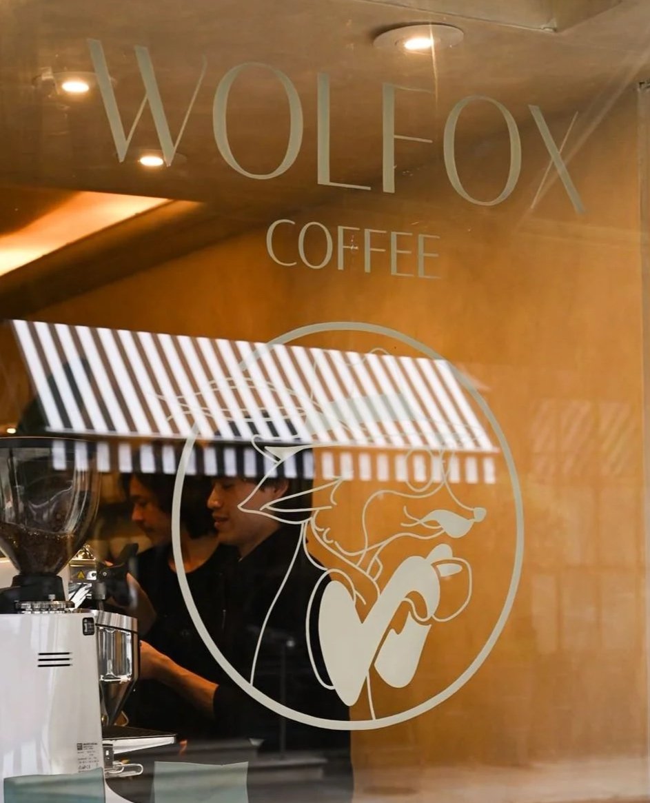 Window of Wolfox Coffee shop with logo of a wolf holding a coffee cup, and two baristas making coffee inside, with Christmas decorations at the bottom. Speciality Coffee, Brunch & Bakery Near Me. London, Lake Como, Brighton