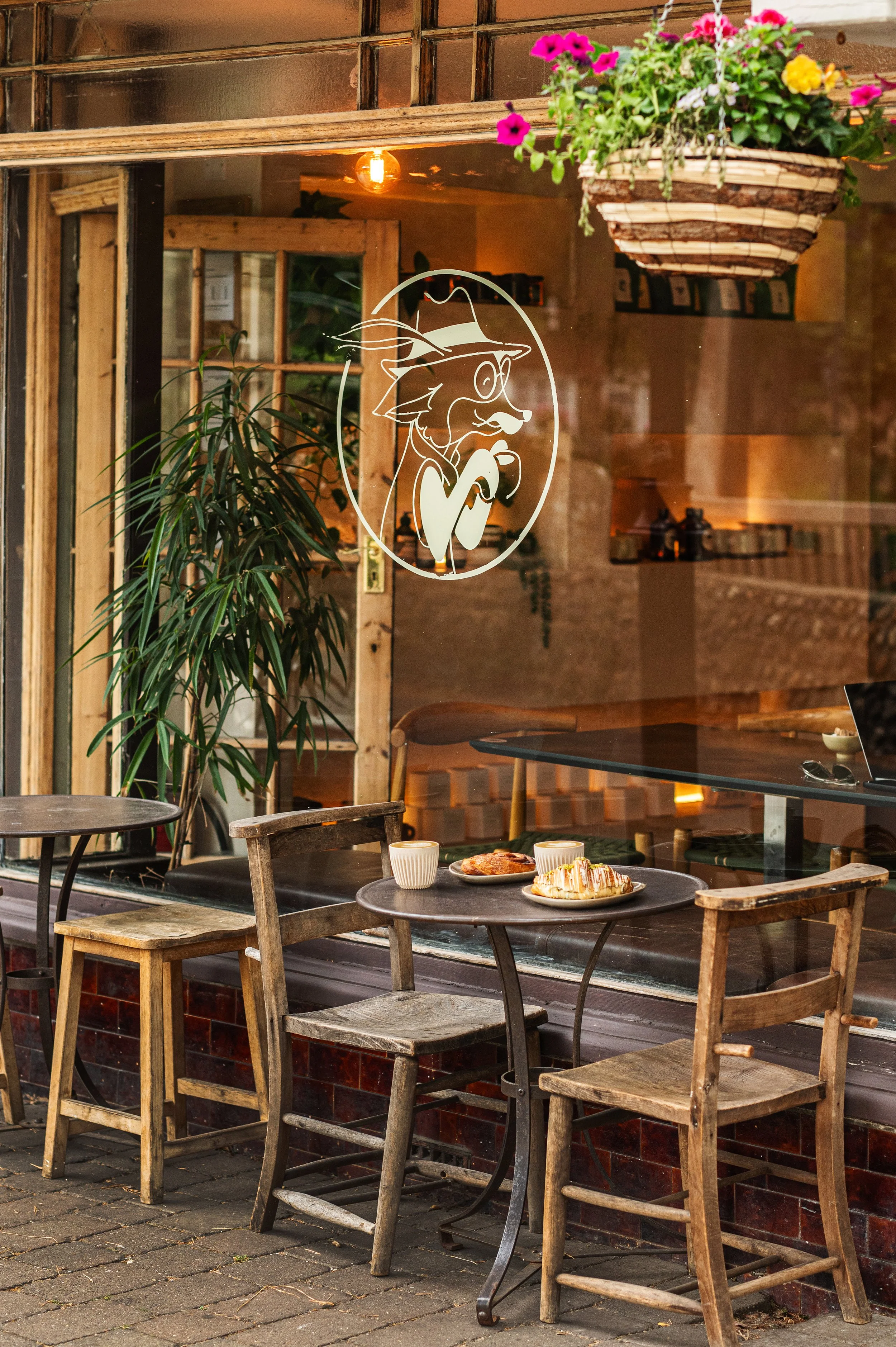 Outside view of a cafe with four wooden chairs and a small round table on a brick sidewalk. The table has a plate of pastries and two cups of coffee. Shorham by sea Sussex - Speciality Coffee, Brunch & Bakery Near Me. London, Lake Como, Brighton