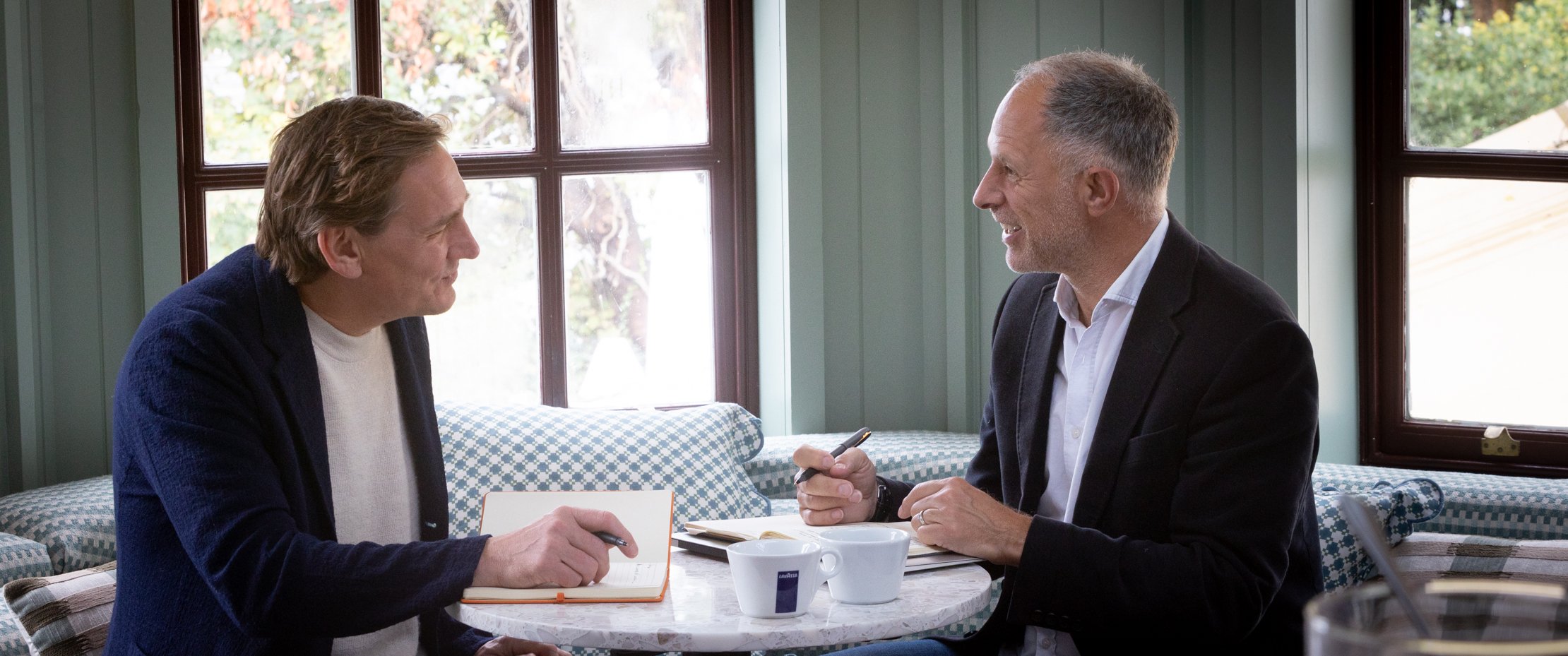 Two men sitting at a table, engaged in conversation, with notebooks and a coffee cup on the table.
