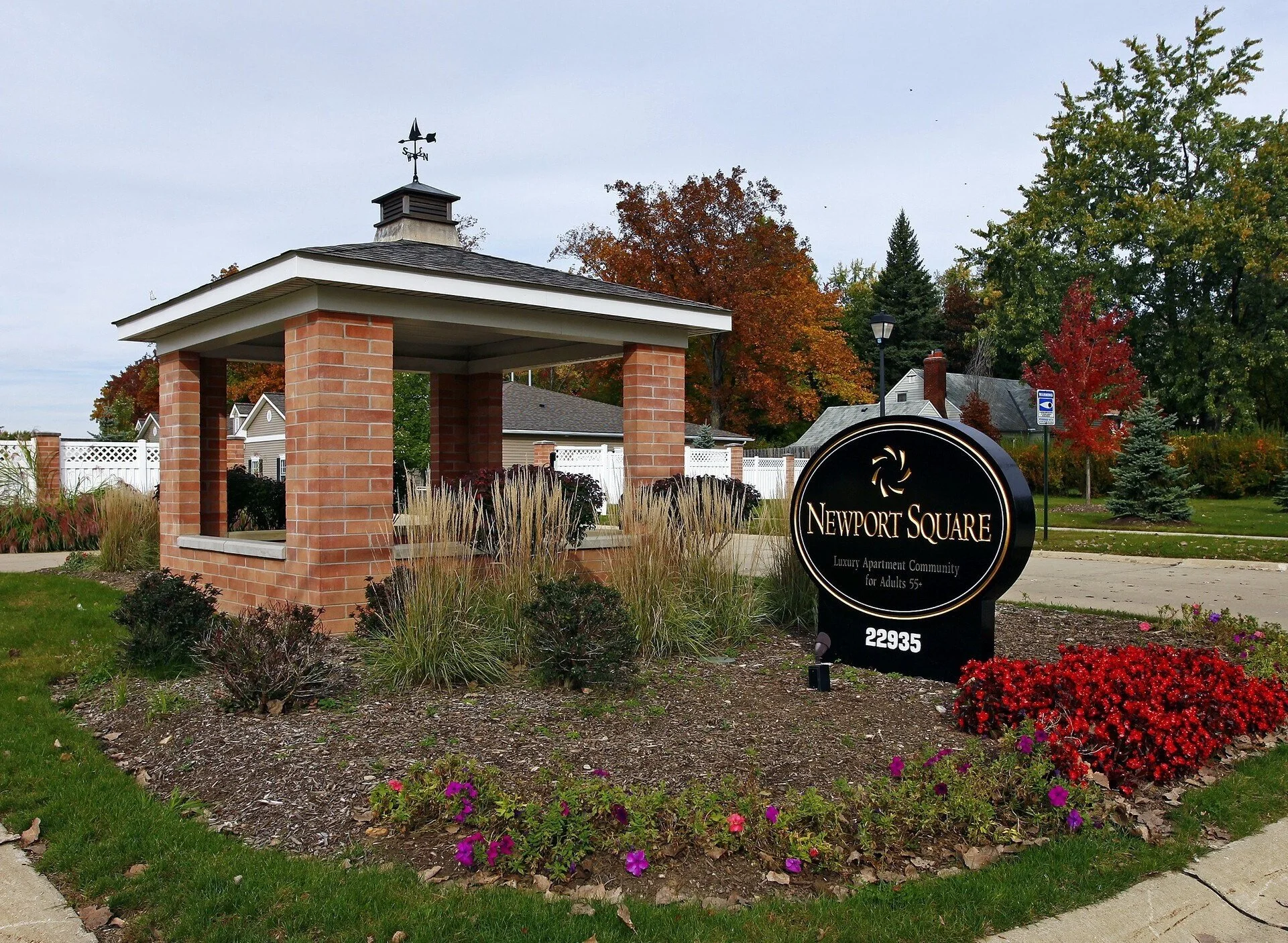 Sign for Newport Square luxury apartment community for adults 55+ at 22935 with a brick shelter and surrounding landscaped plants and flowers in a suburban area.