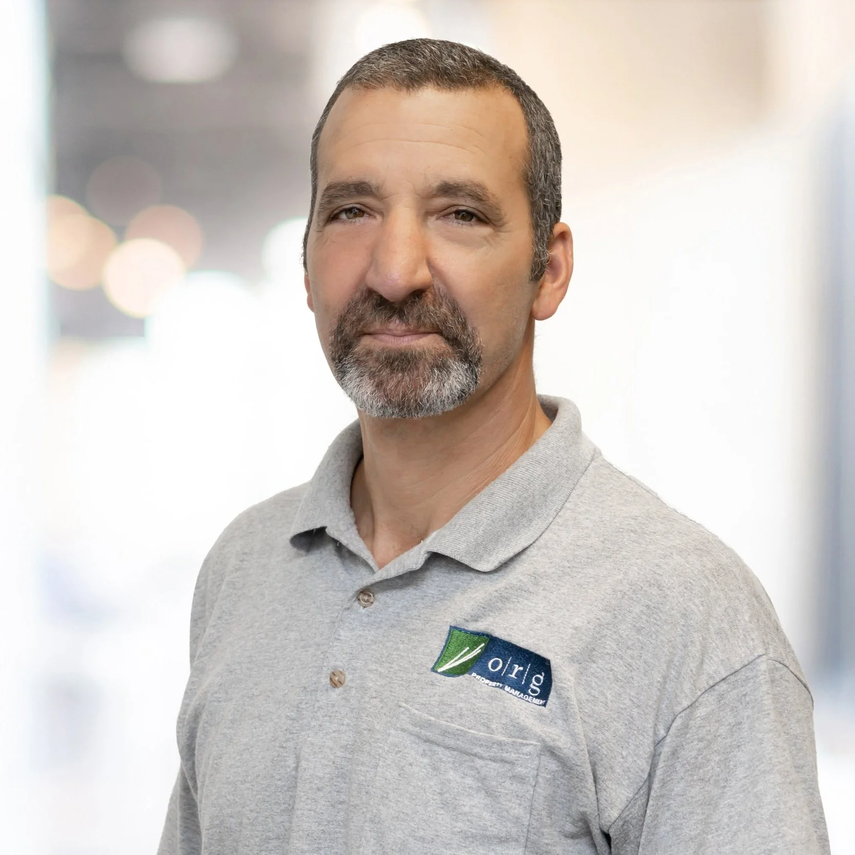 Middle-aged man with short hair and a beard, wearing a light gray polo shirt with an 'org' logo, standing indoors with blurred background.