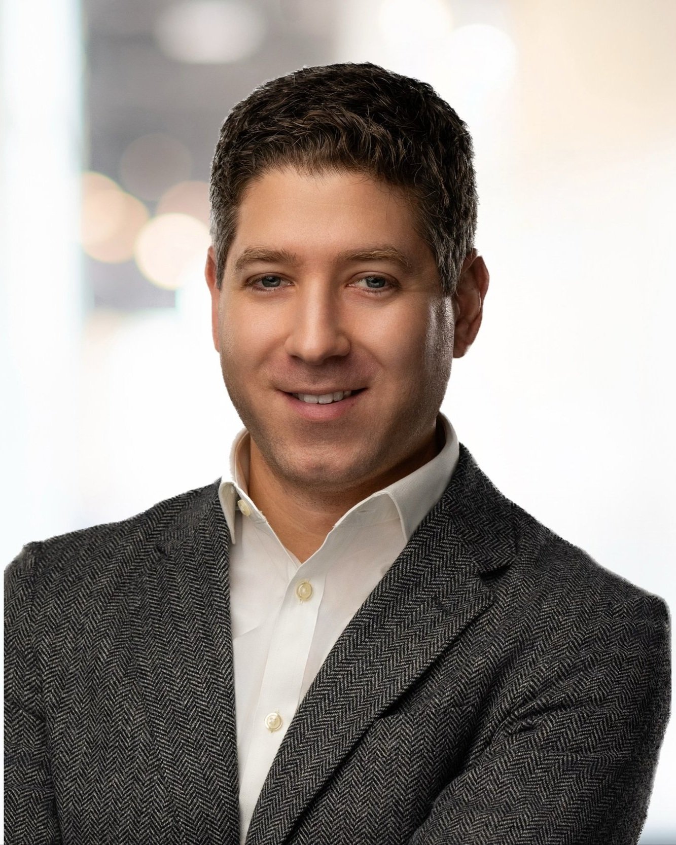 A young man in a white shirt and gray blazer smiling confidently in a professional indoor setting.