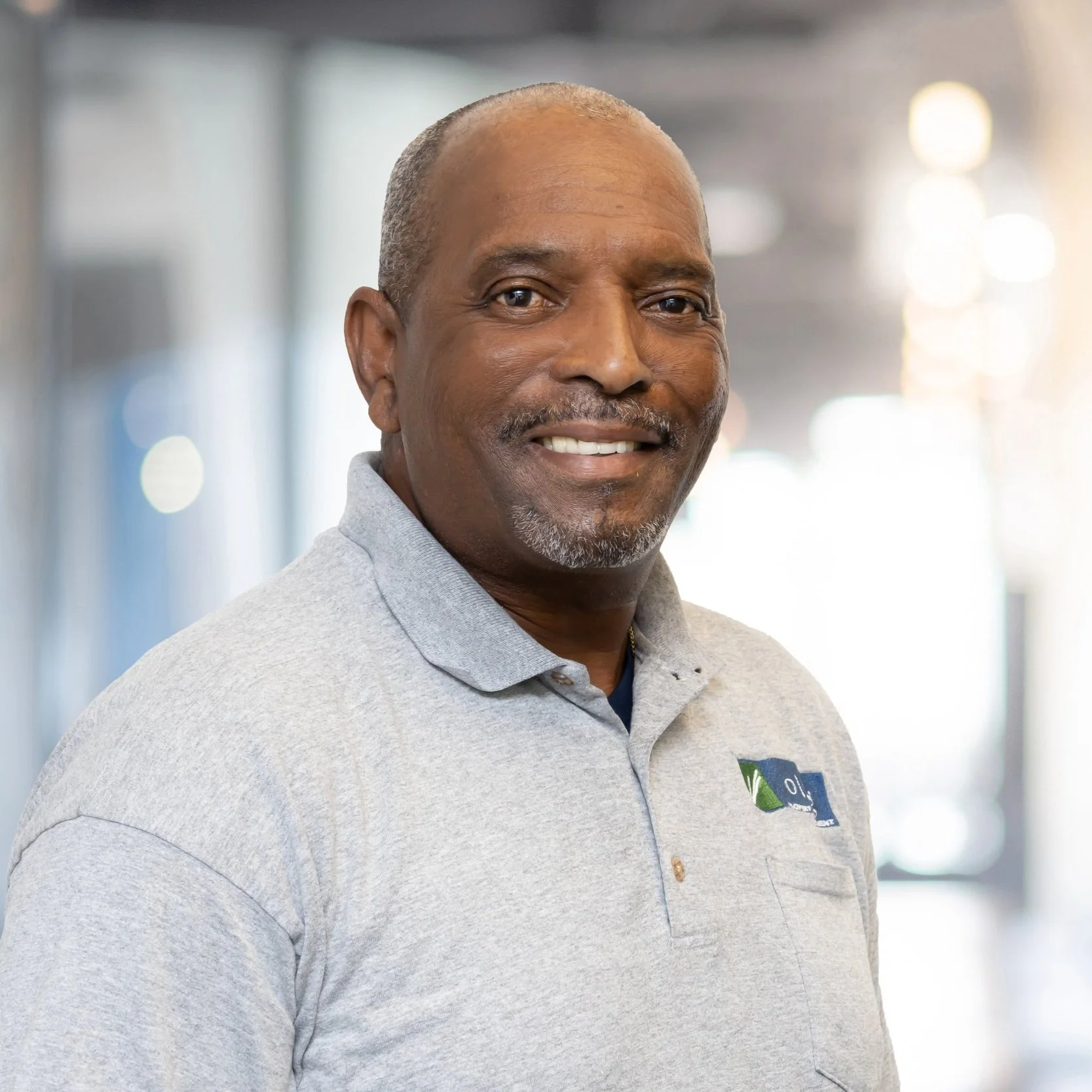 A middle-aged man with short gray hair and a goatee, smiling, wearing a light gray polo shirt with a logo on the left chest, standing inside a well-lit building with blurred background lights.