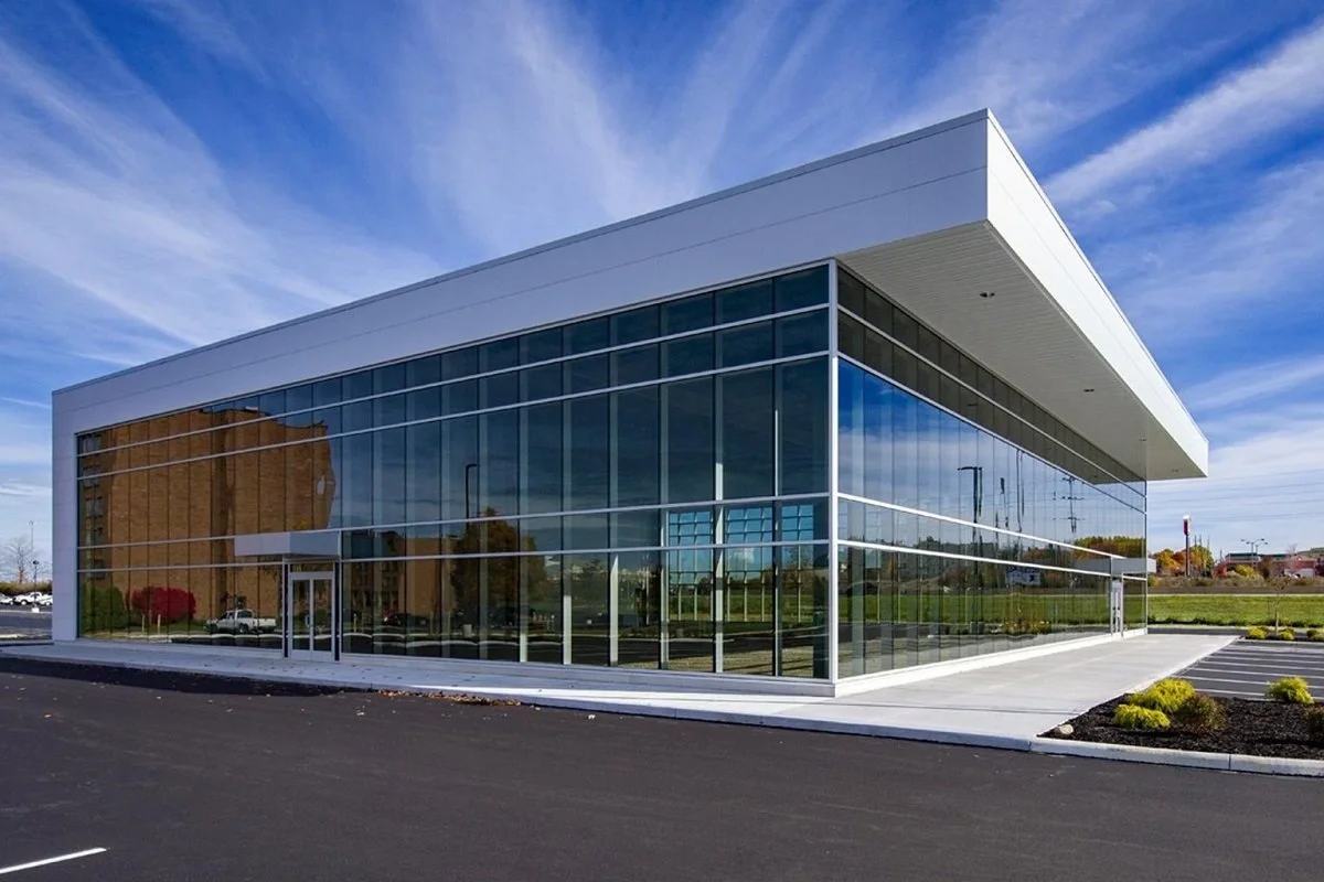 Modern glass commercial building with a white overhang, surrounded by a parking lot and landscaped areas, under a blue sky with wispy clouds.