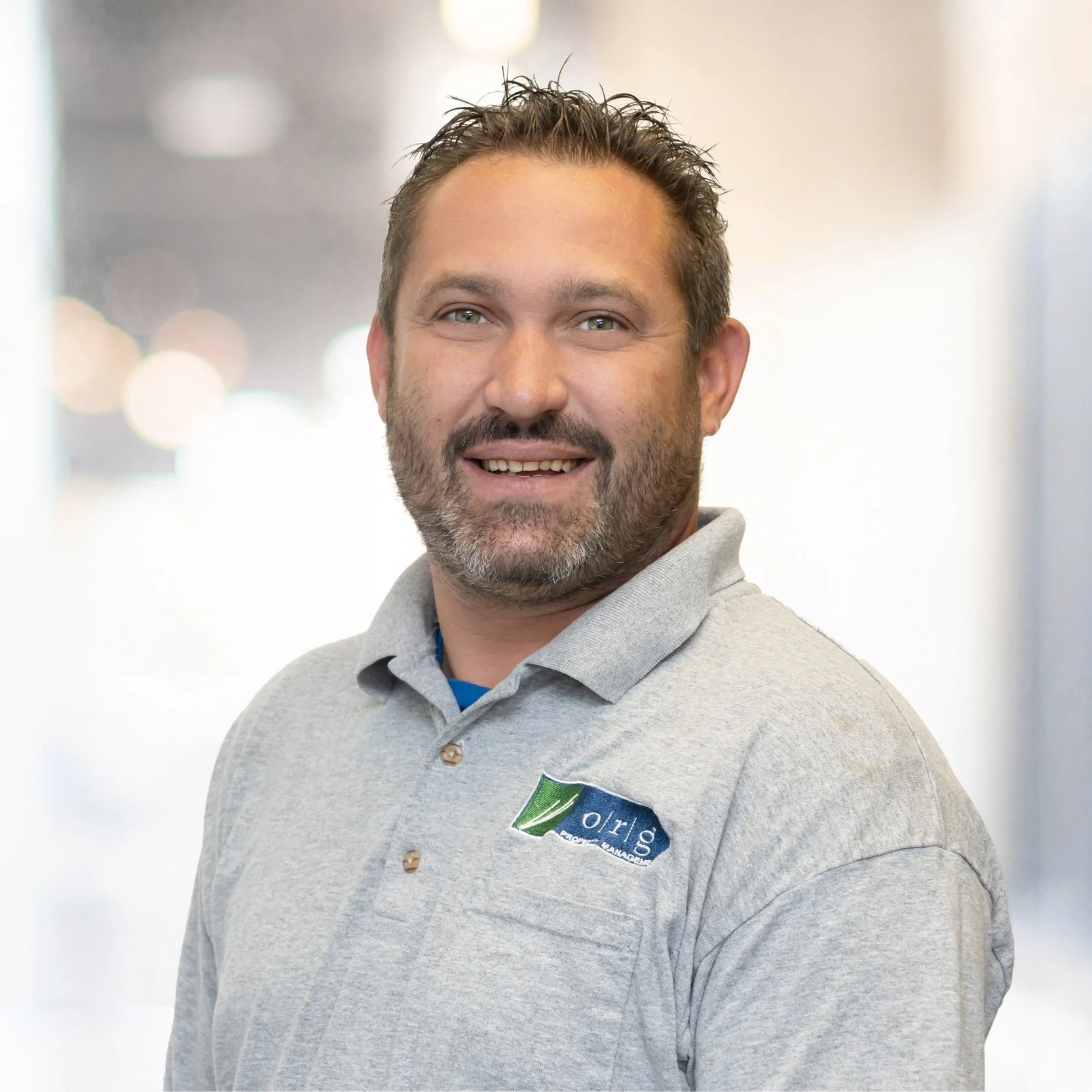 A man with short, spiky hair and a beard, smiling, wearing a light gray polo shirt with a logo on the chest that reads 'orlg'. The background is blurred with bright bokeh lights.
