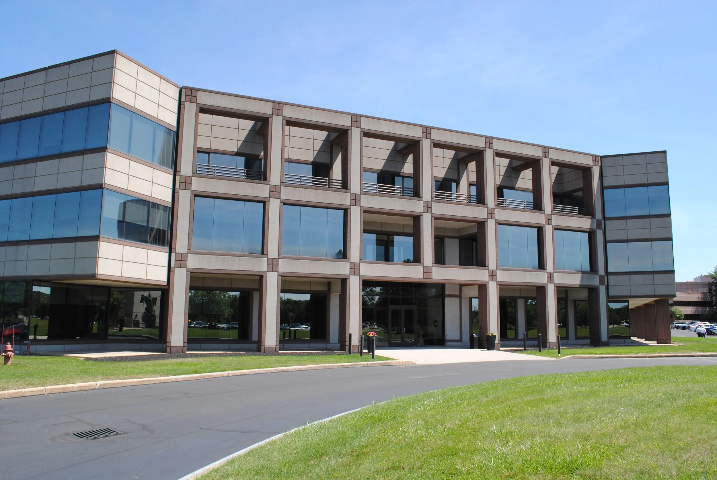 Modern office building with large glass windows, surrounded by green lawn and a paved driveway, under a clear blue sky.