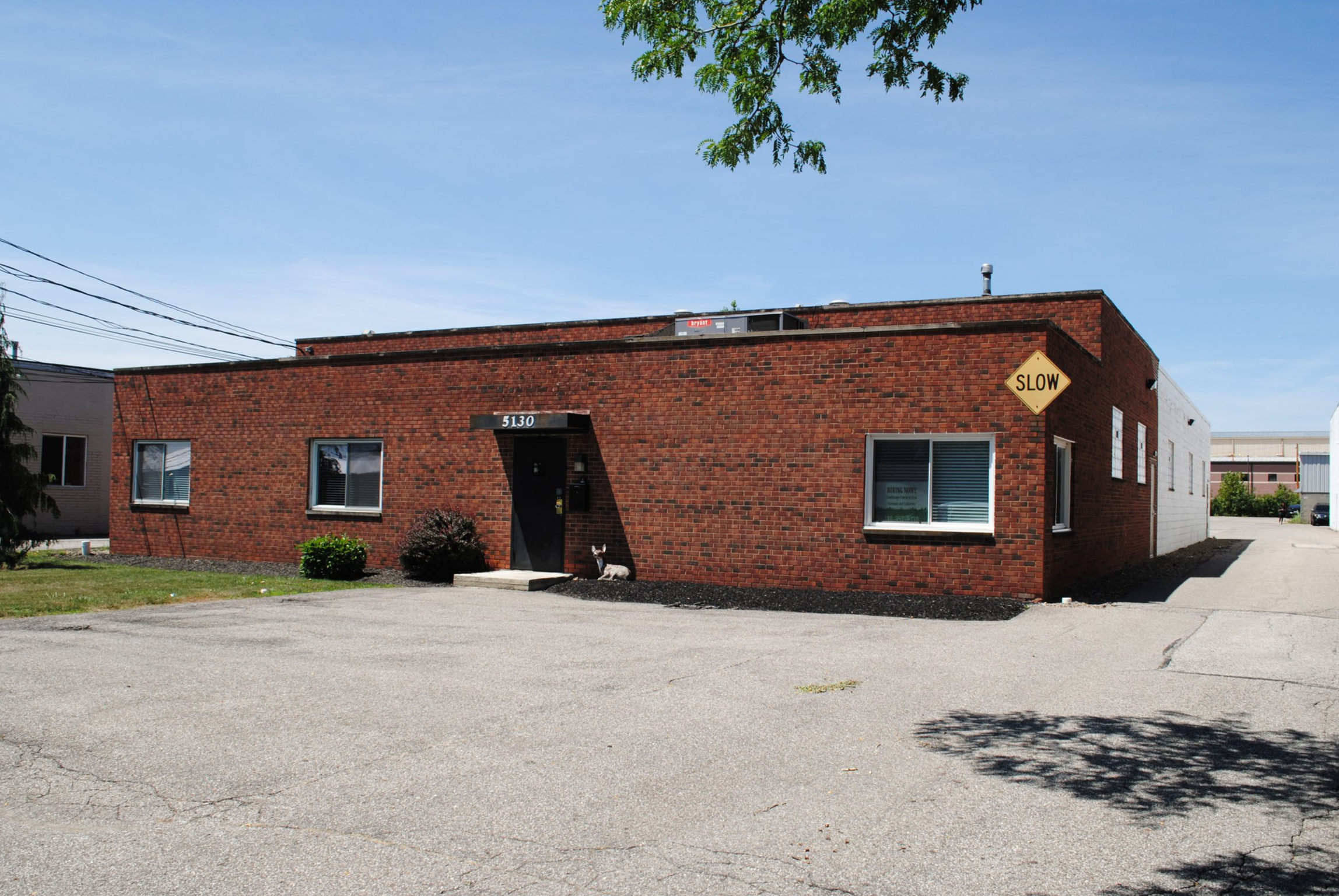 A single-story brick building with three windows on the front, a black door, and the number 5130 above the door. There is a sign that says 'SLOW' on the corner of the building. In front, there is a parking lot with visible cracks and a small dog sitting near the door, and some landscaping with small bushes and mulch.