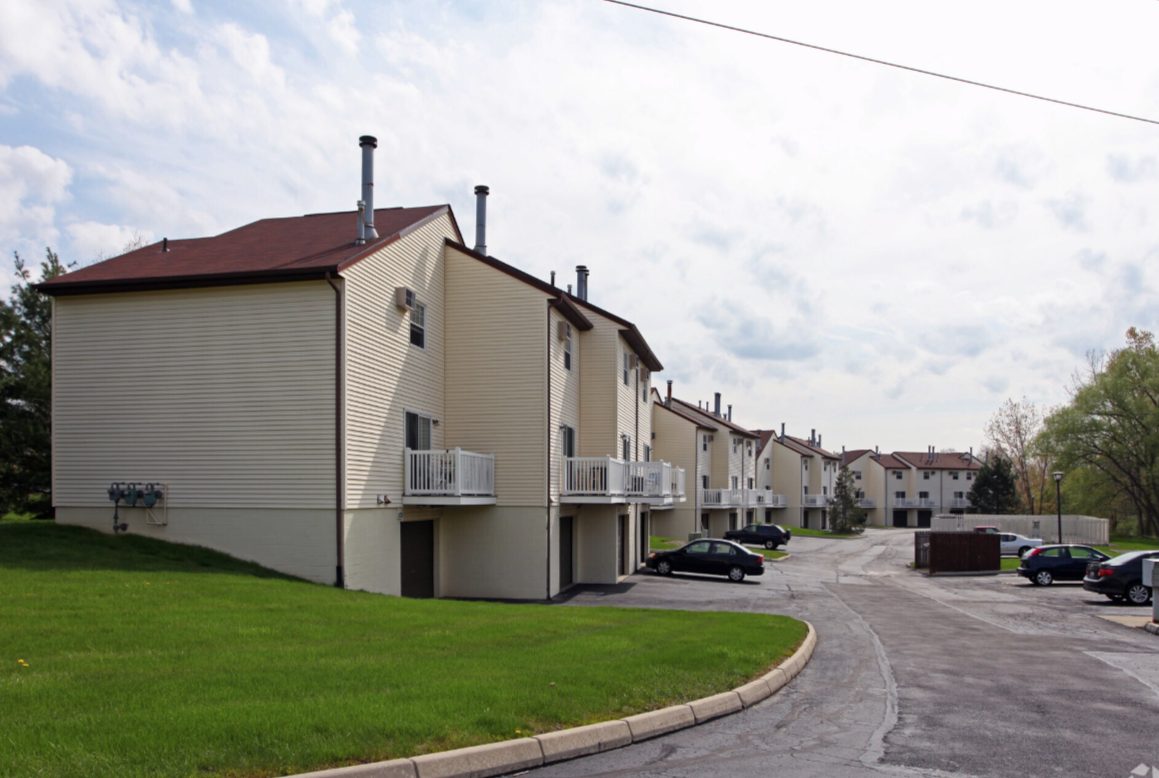 A row of beige multi-story townhouses with small balconies, asphalt parking lot with parked cars, green lawn, and partly cloudy skies.