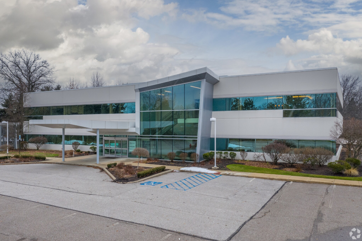 Modern office building with large glass windows, surrounded by a parking lot, some older trees, and cloudy sky.