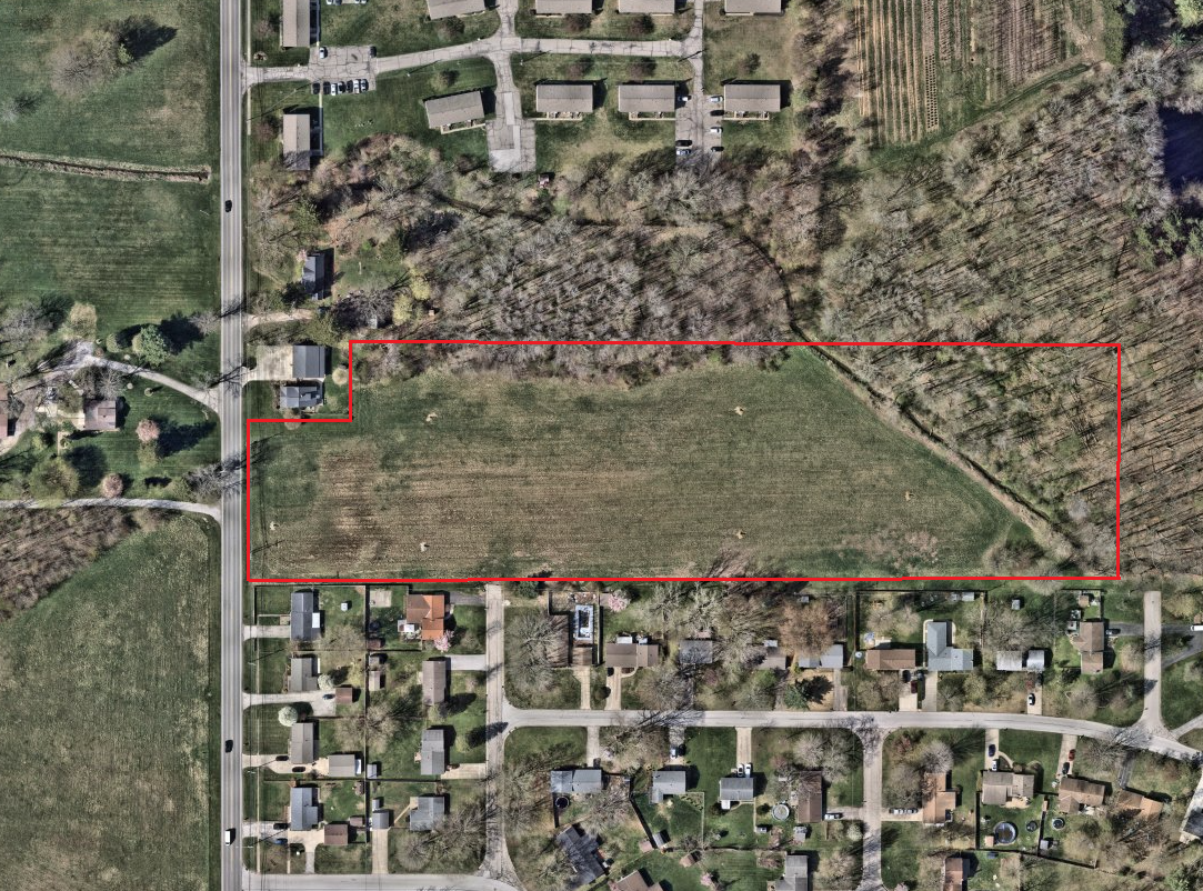 A large rectangular grassy field outlined in red, surrounded by residential houses, trees, and a road.