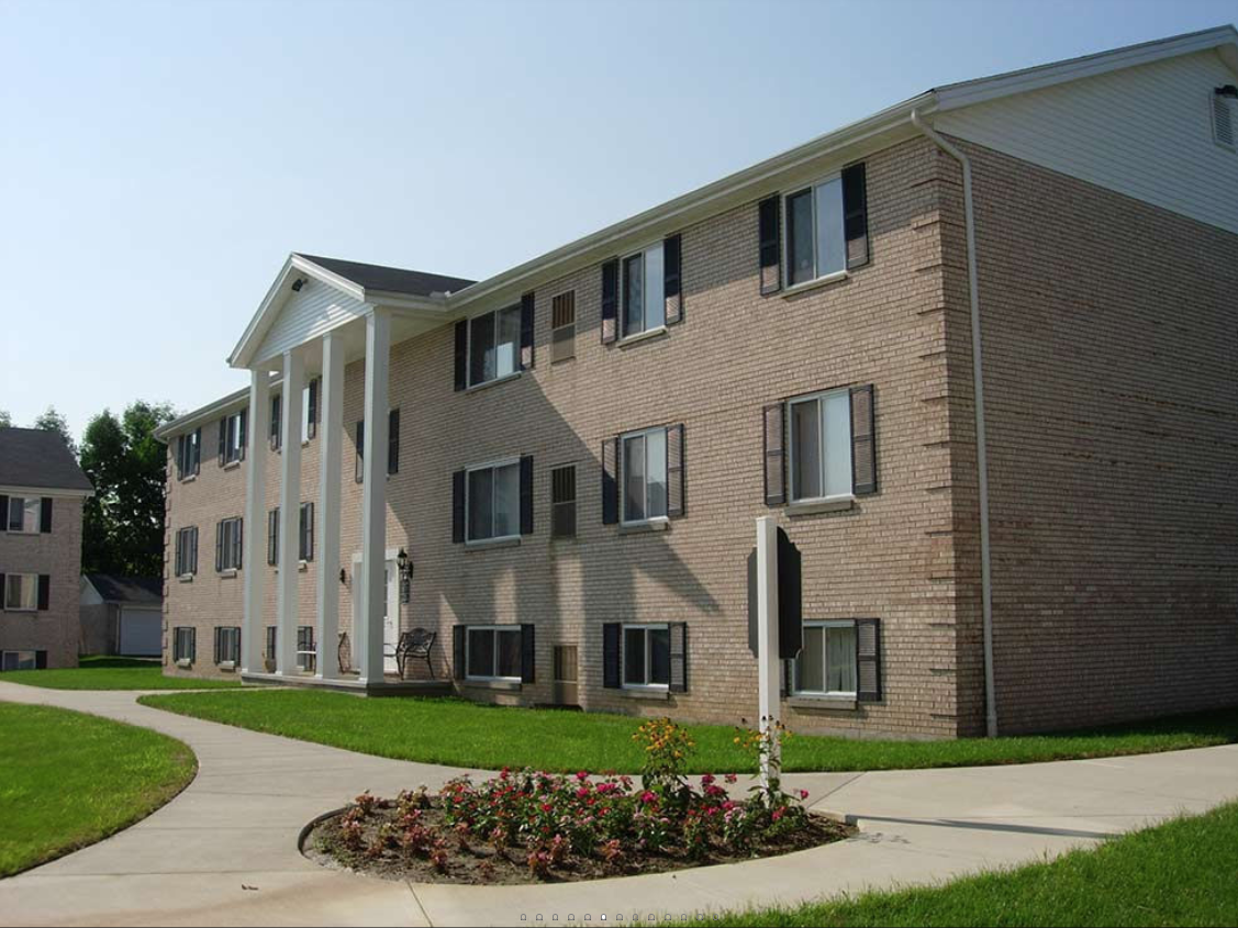 Three-story brick apartment building with black shutters, white columns at entrance, sidewalk, green lawn, and flower bed in foreground under clear blue sky.