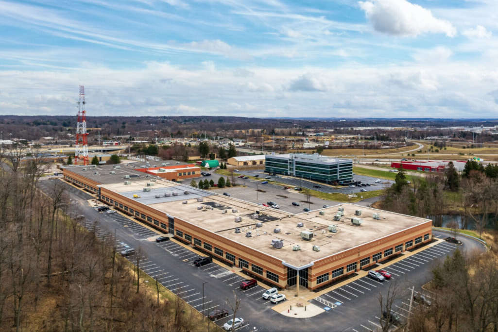 An aerial view of a large commercial building with a flat roof, surrounded by parking lots, with trees in the foreground and a highway in the background under a partly cloudy sky.