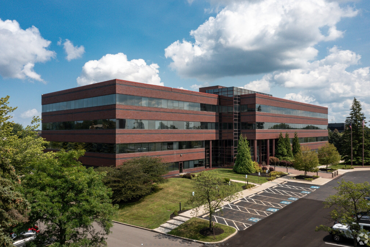 A multi-story office building with a brick exterior and large windows, surrounded by parking spaces, trees, and green landscaping under a partly cloudy sky.