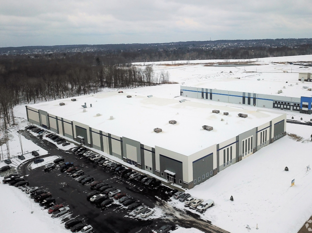 An aerial view of a large warehouse building with a snow-covered roof, surrounded by a parking lot with multiple cars, with a snow-covered field and trees in the background during winter.
