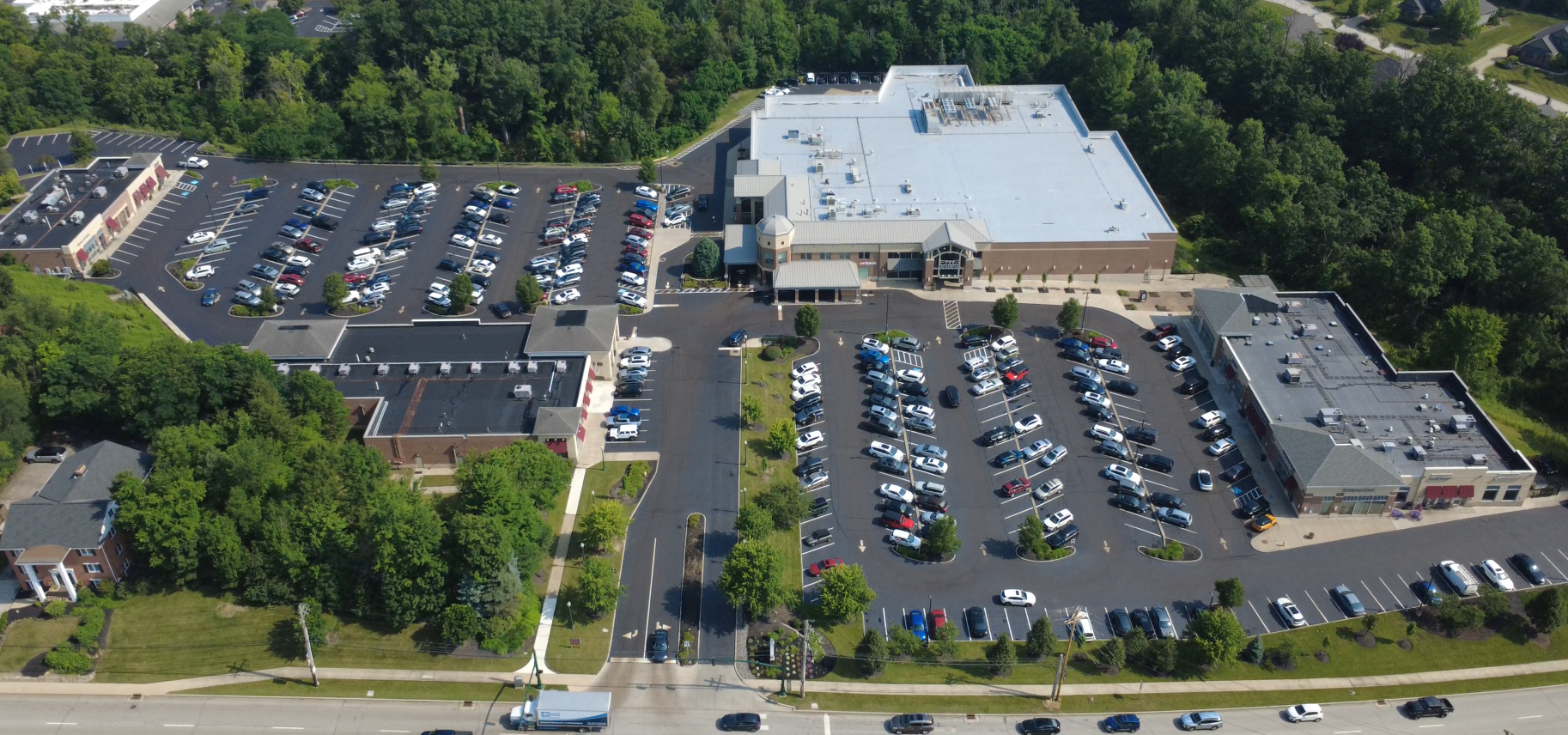 Aerial view of a shopping mall parking lot with multiple cars parked, surrounded by trees and buildings.