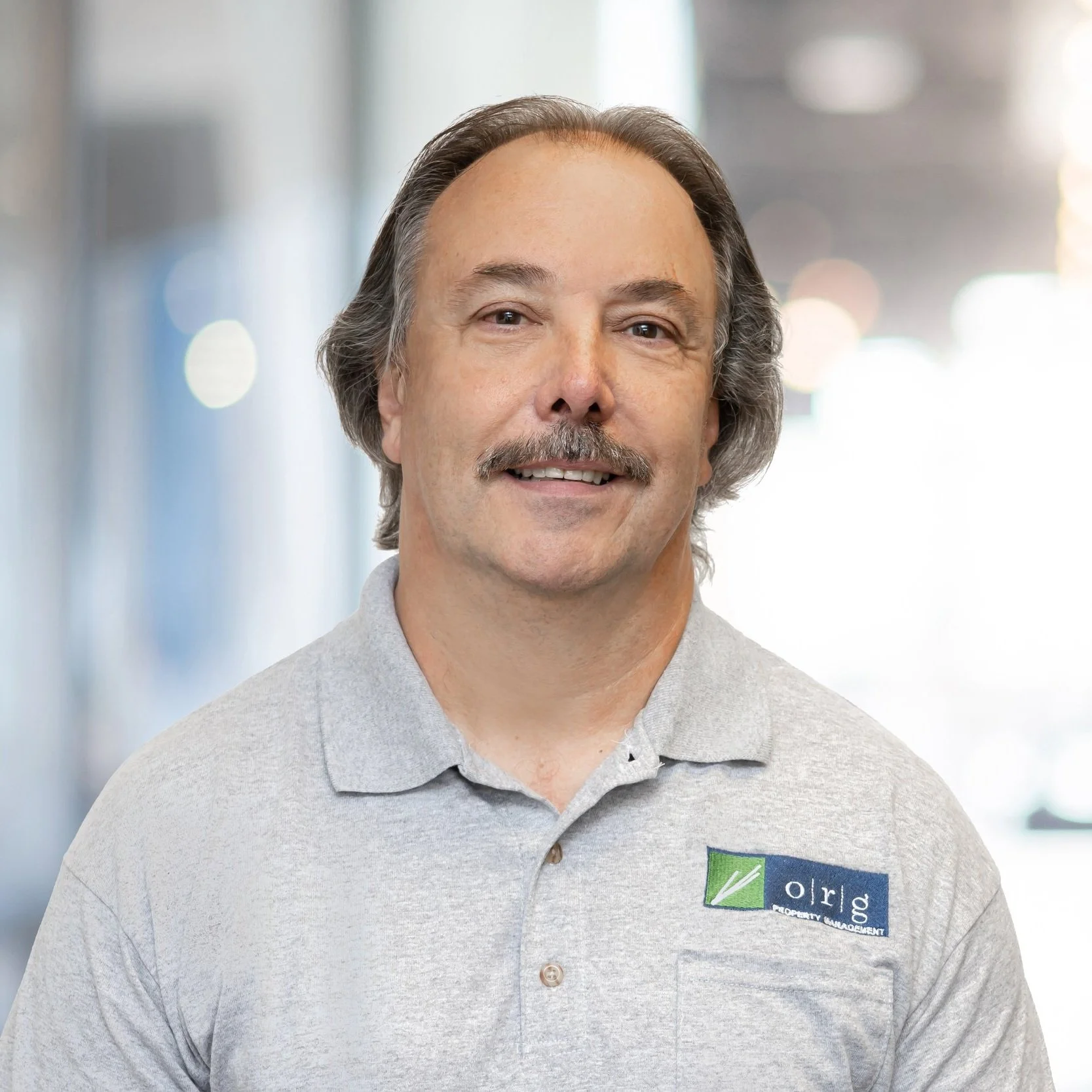 A middle-aged man with gray hair, a mustache, and a goatee, wearing a light gray polo shirt with a company logo, smiling at the camera. The background is blurred indoor setting.