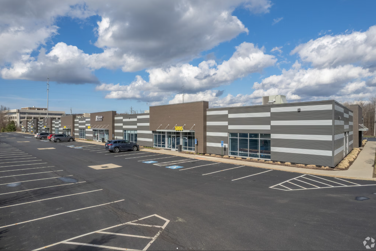 Commercial strip mall with multiple storefronts, parking lot with several cars, and handicap parking spaces, under a partly cloudy sky.