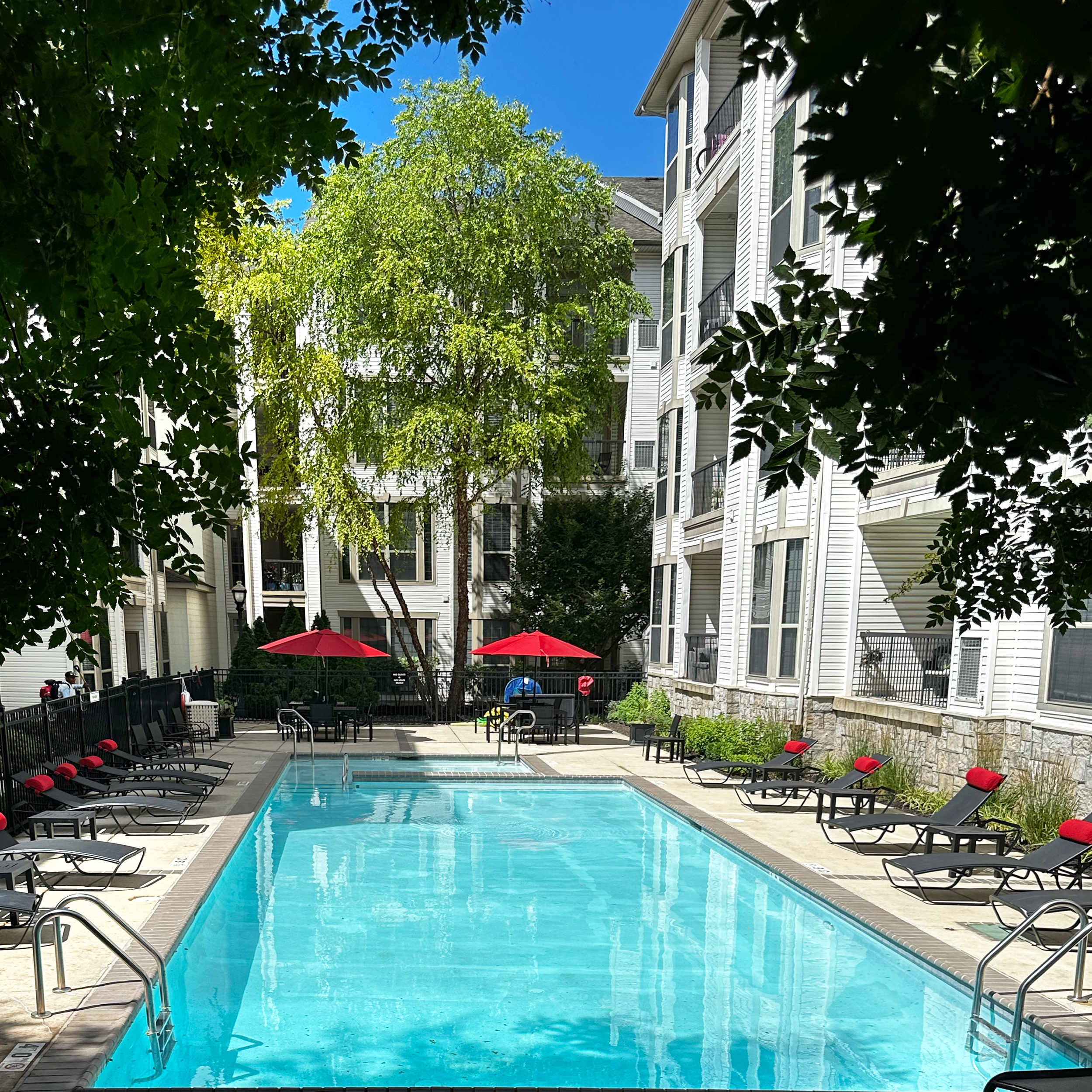 Outdoor swimming pool with lounge chairs and red umbrellas, surrounded by apartment buildings and trees.