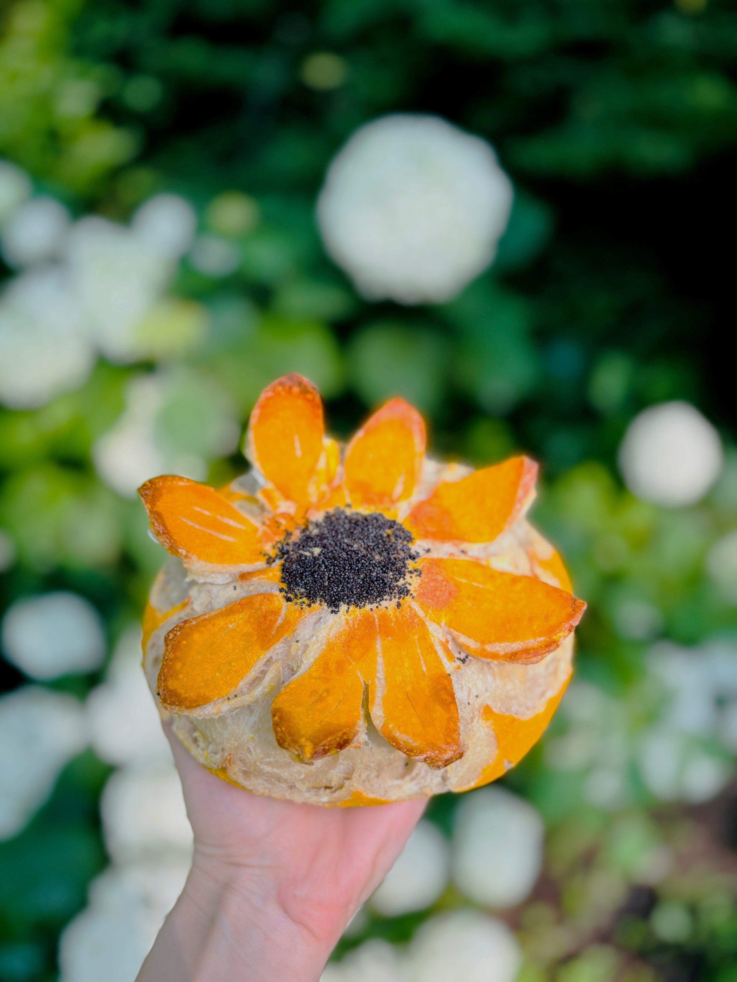 Hand holding a sourdough bread loaf scored in petal-like sections, resembling a flower, with a black seed-filled center, in front of a garden with white flowers.