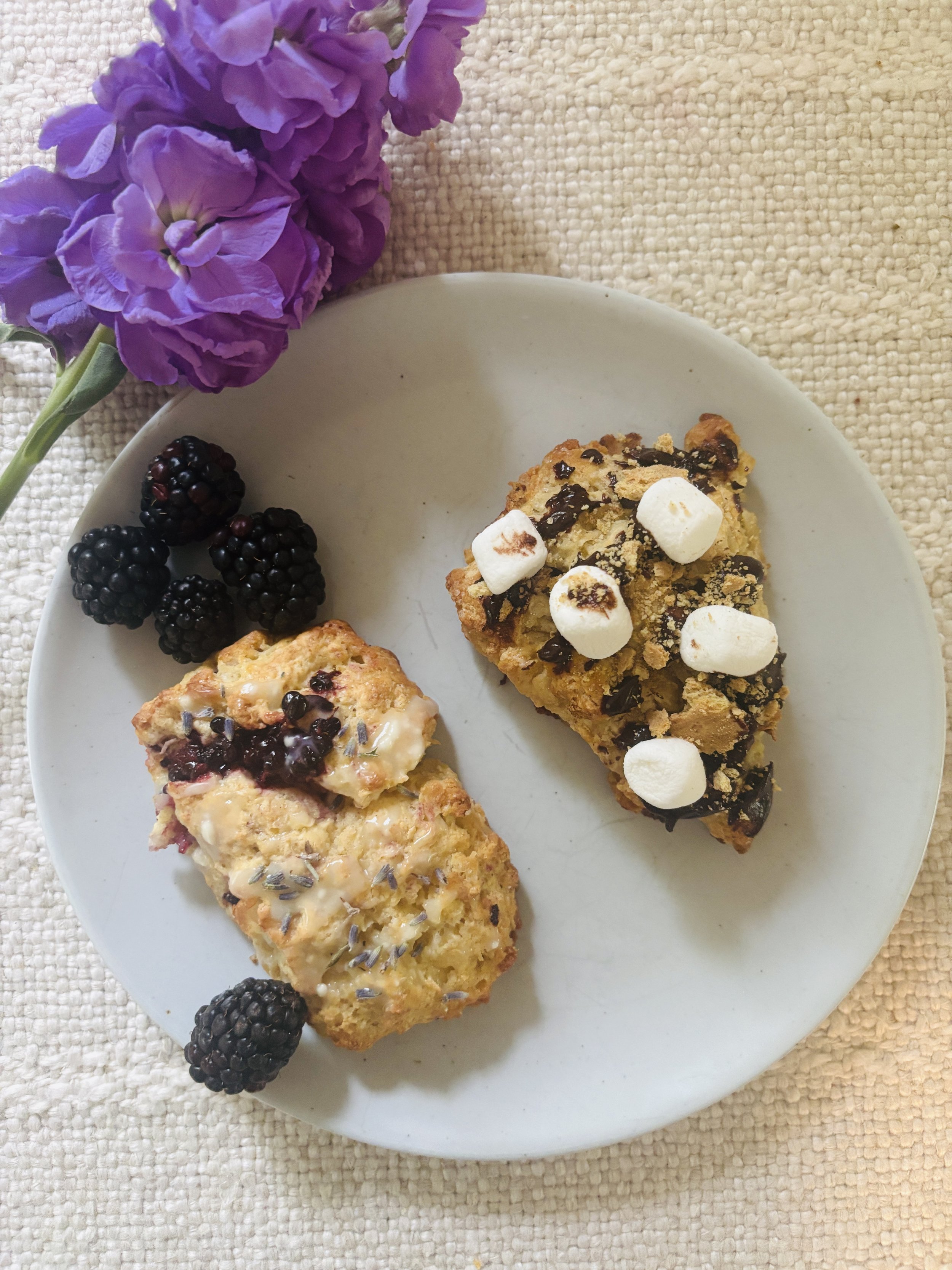 A white plate with two sourdough scones alongside fresh blackberries and a purple flower on a textured beige surface.