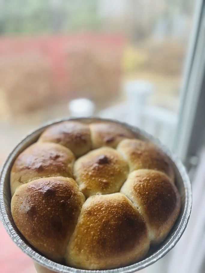 Close-up of a tray of freshly baked dinner rolls with a golden-brown crust, held near a window with a blurred outdoor background.