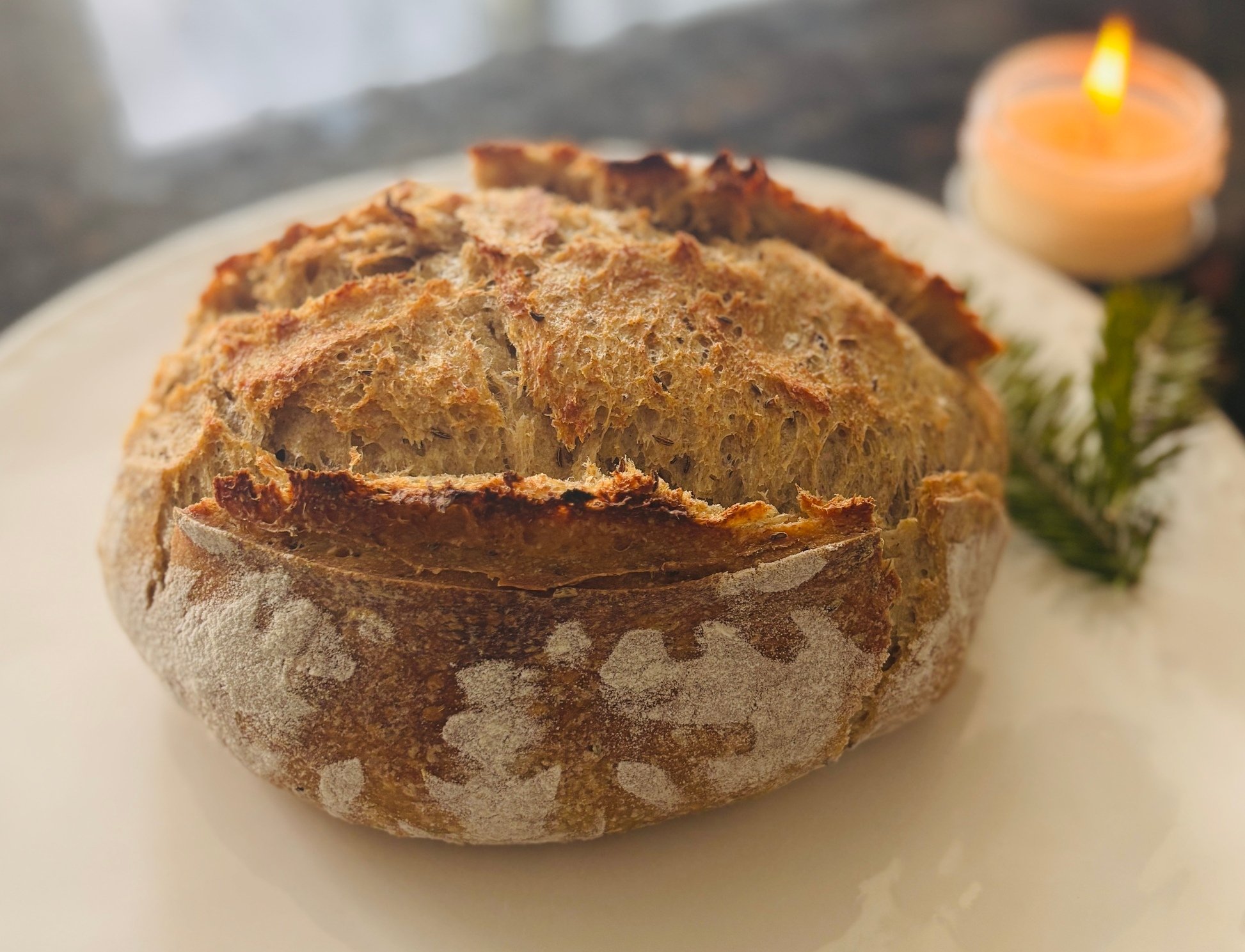 Close-up of a rustic loaf of bread on a white plate with a lit candle and greenery in the background.