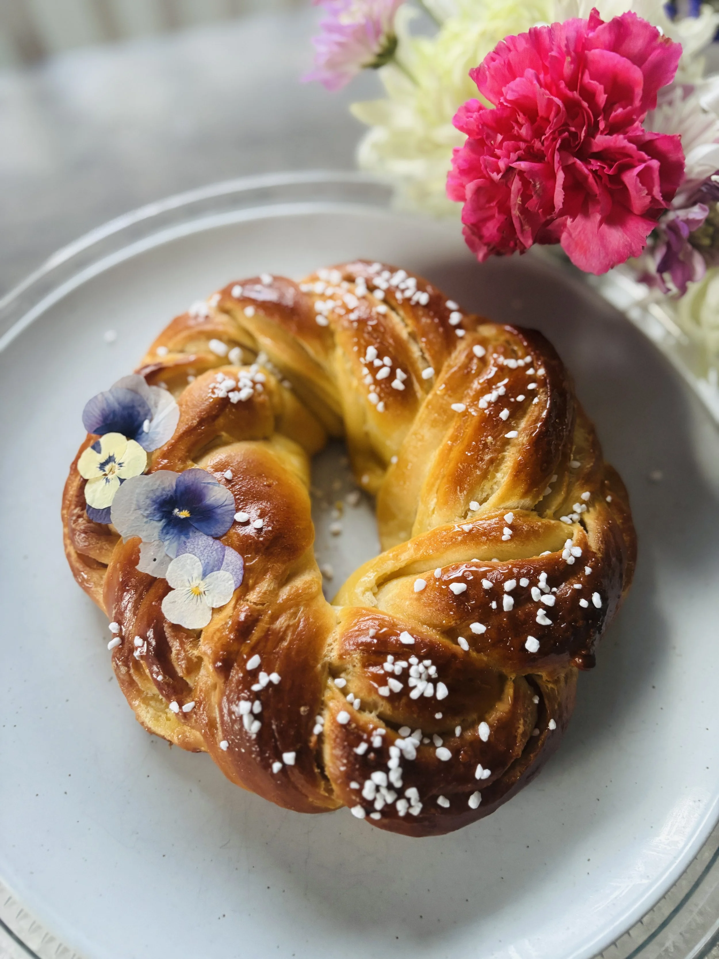 A round pastry with a braided pattern, topped with pearl sugar, and decorated with edible flowers, placed on a white plate.