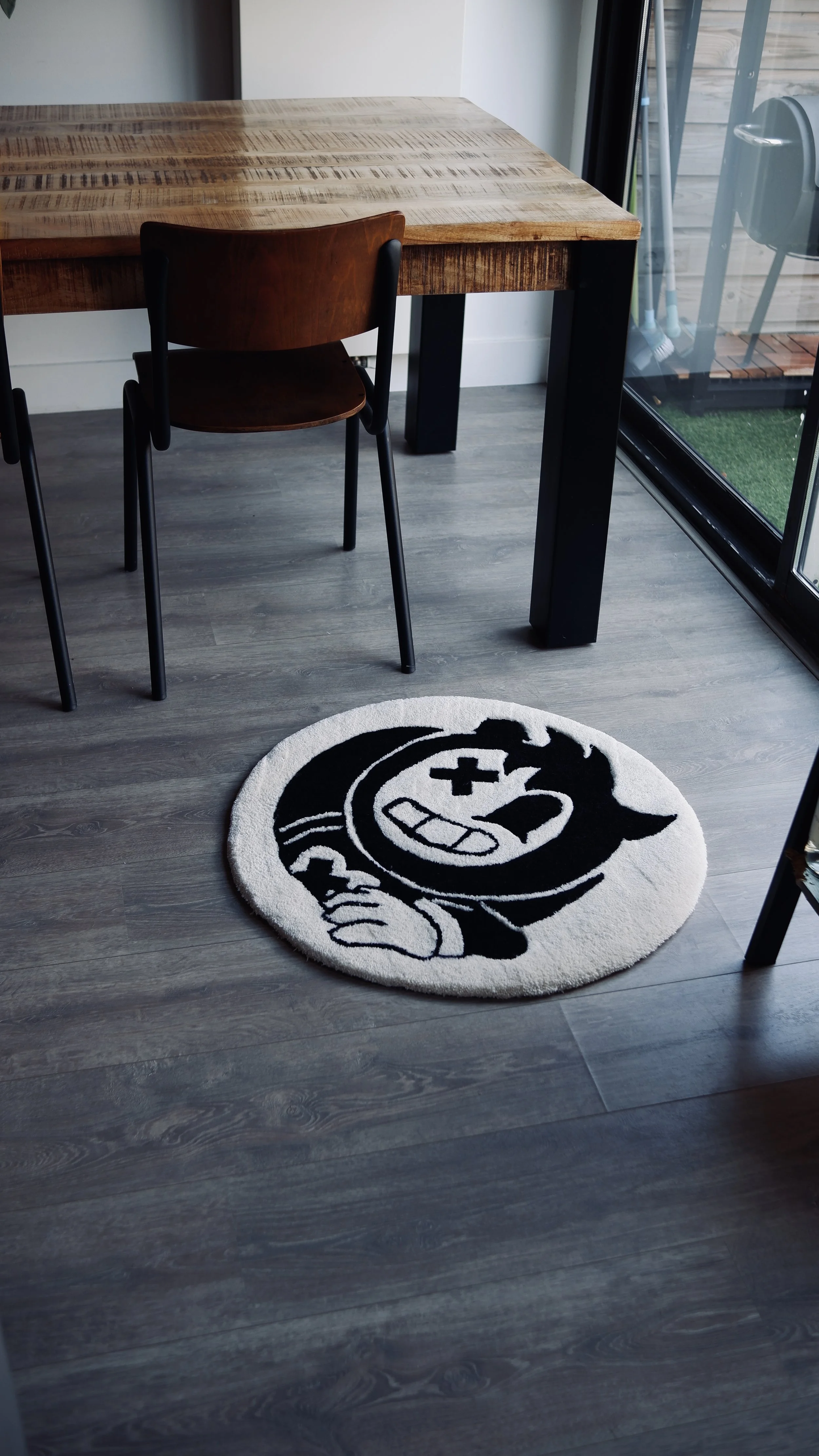A round black and white rug featuring a cartoon character with a big grin, crossed eyes, and a pointing hand, placed on a gray wood floor near a wooden dining table and brown chair, with a sliding glass door and outdoor space visible in the background.