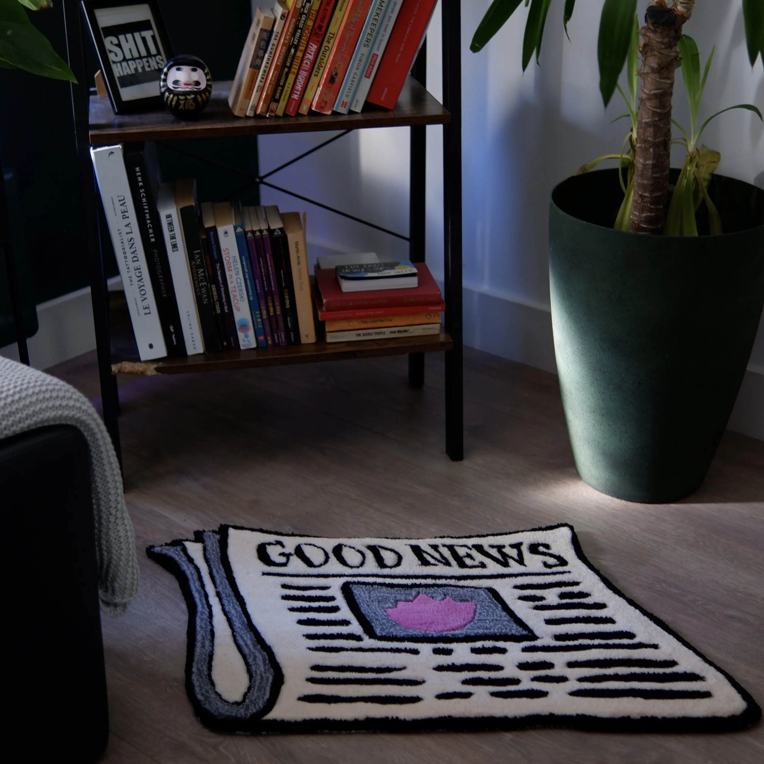 Living room with a black bookshelf filled with books, a small framed picture with text that says 'SHIT HAPPENS,' a decorative item, and a small black and white ornament. A large green potted plant is next to the bookshelf. On the floor, there is a white and black rug with the words 'GOOD NEWS' and an illustration of a pink fish.