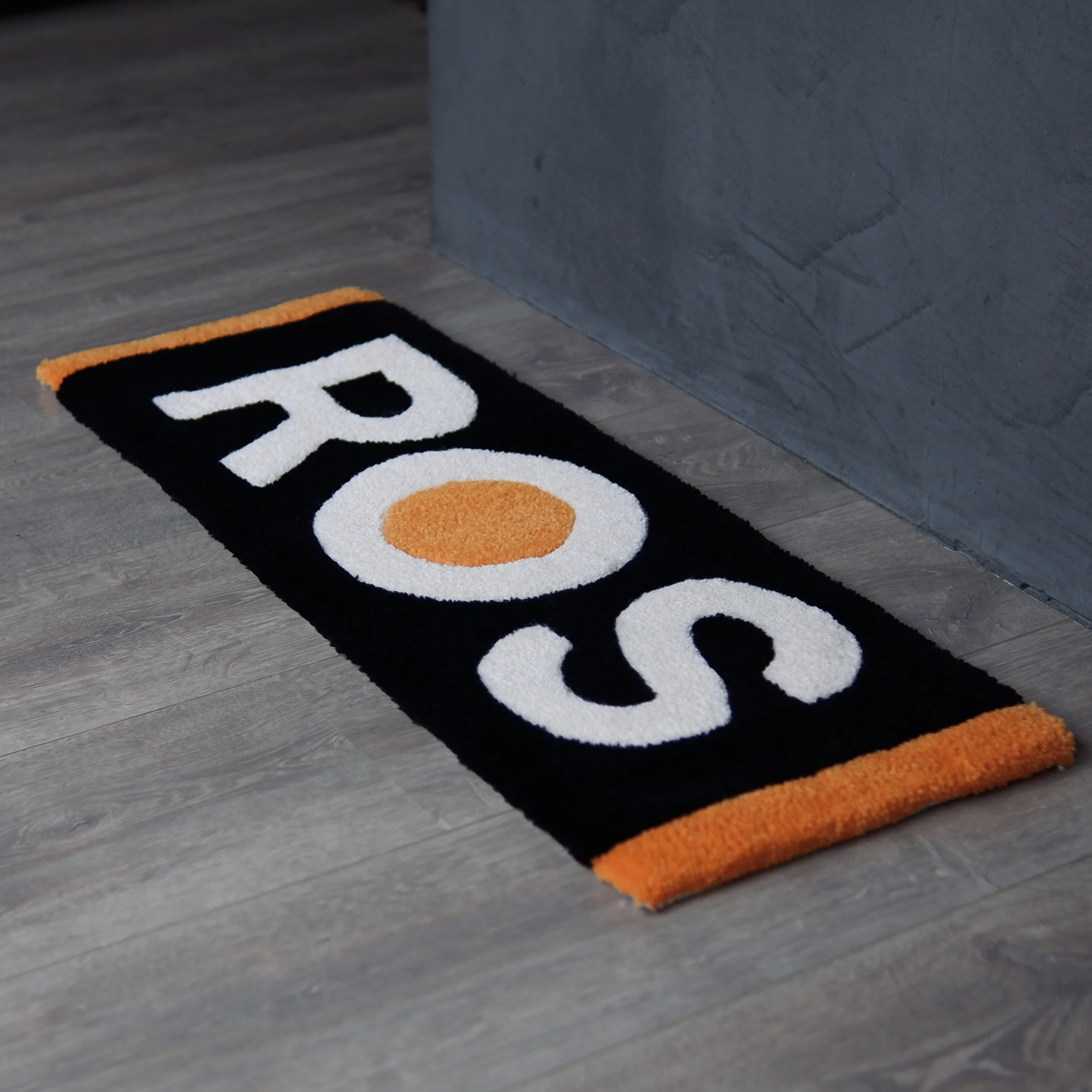 A rectangular doormat with the word 'ROS' in white and orange on a black background, placed on a wooden floor near a gray wall. It's a business logo rug commission piece. 
