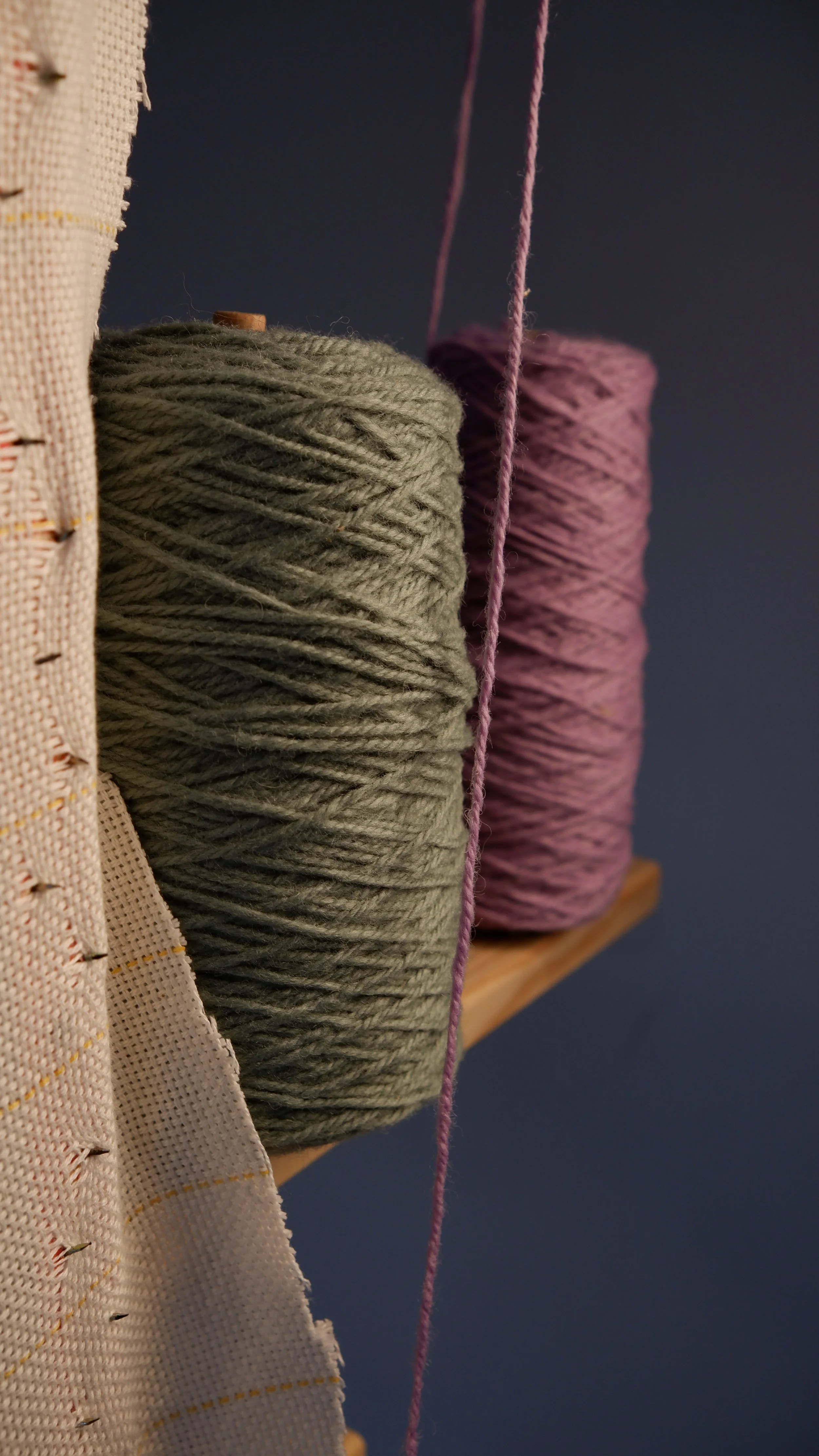 Close-up of two large balls of yarn, one gray and one purple, on a wooden shelf against a dark blue wall, with a piece of fabric and a pink strand of yarn hanging in the foreground.