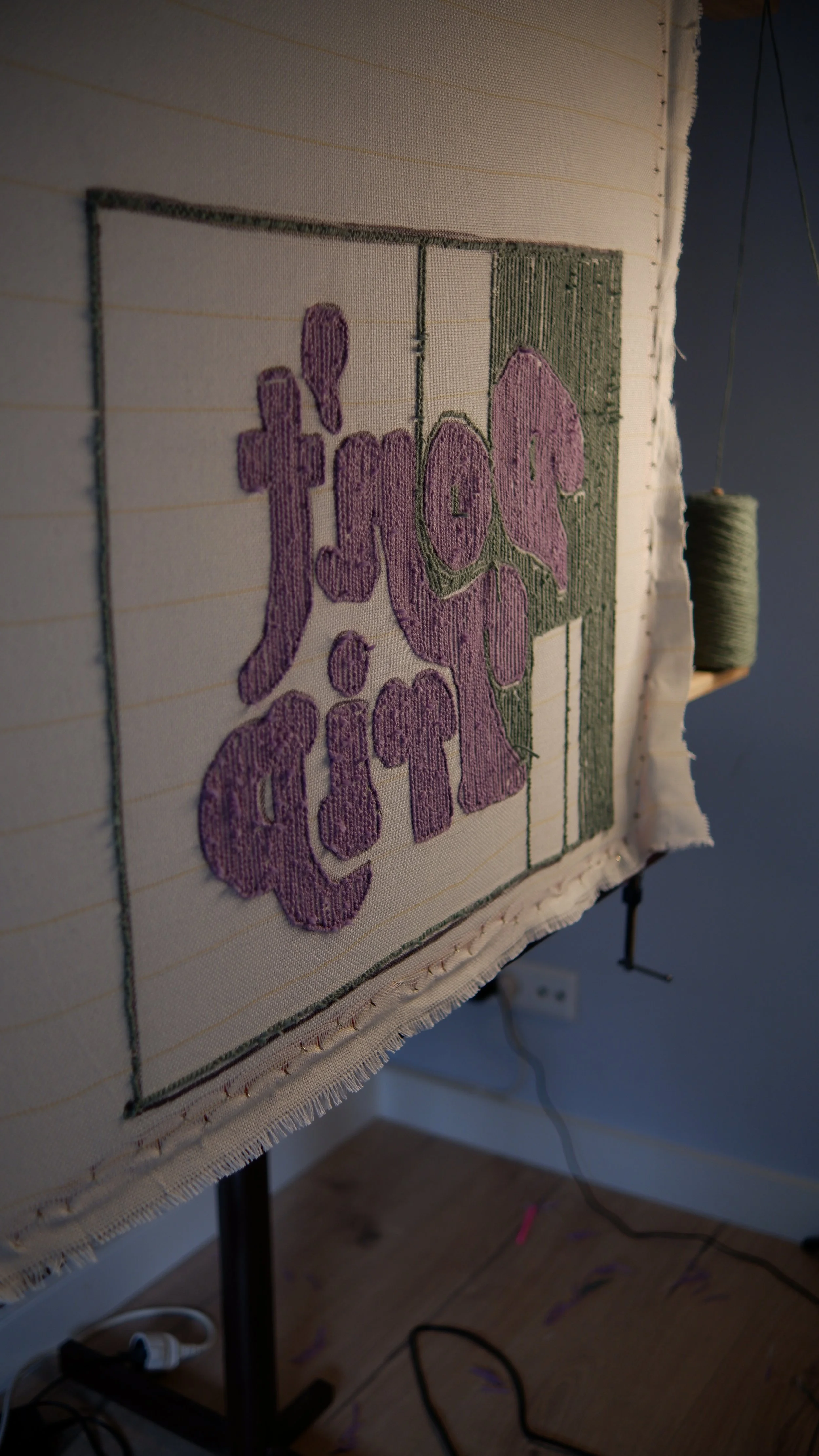 Close-up view of an embroidery hoop holding a piece of fabric with a stitched pattern of purple and green text, with a spool of green thread hanging in the background.