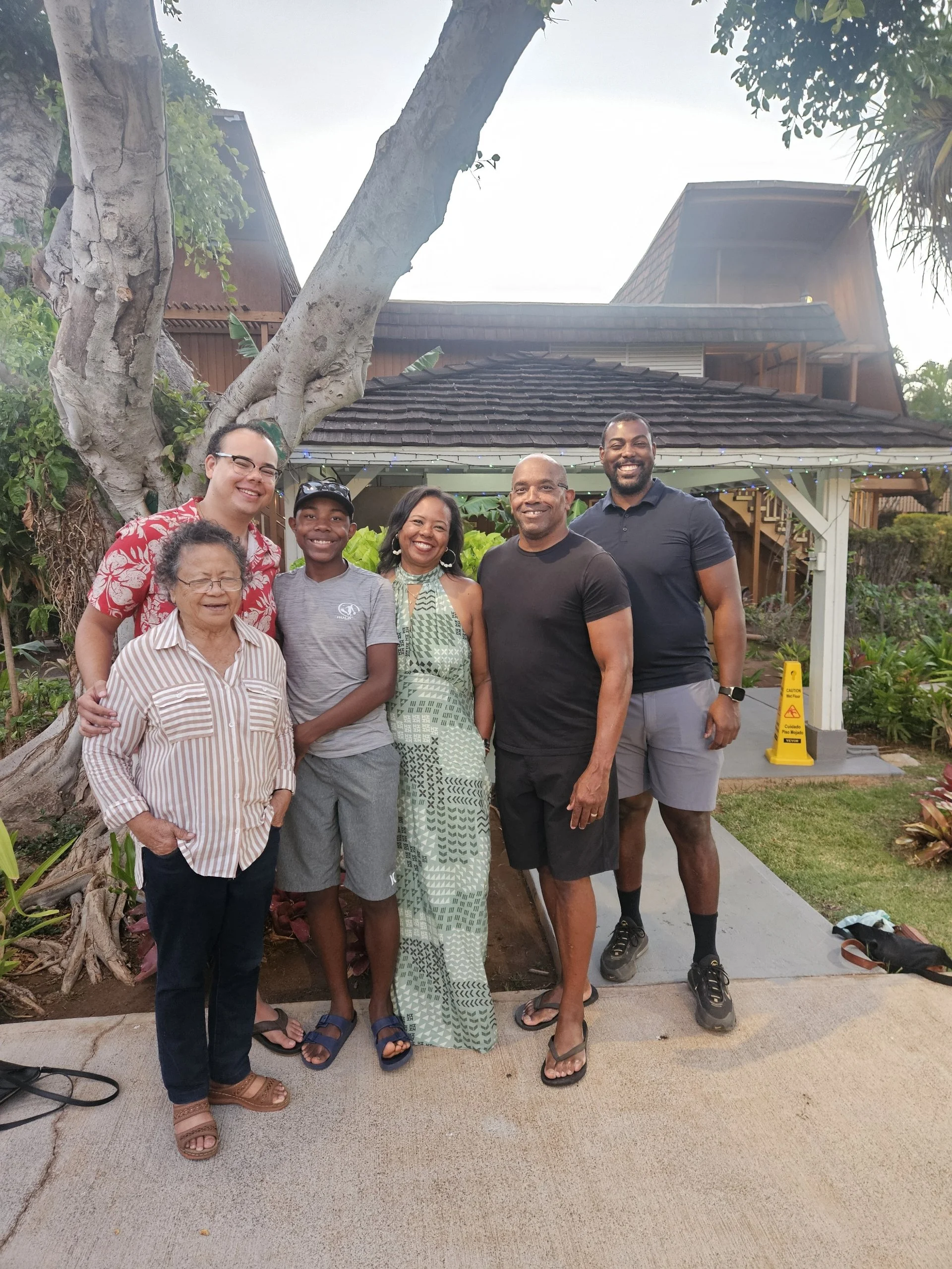 Group of seven diverse people smiling and posing outdoors in front of a house with trees and greenery.