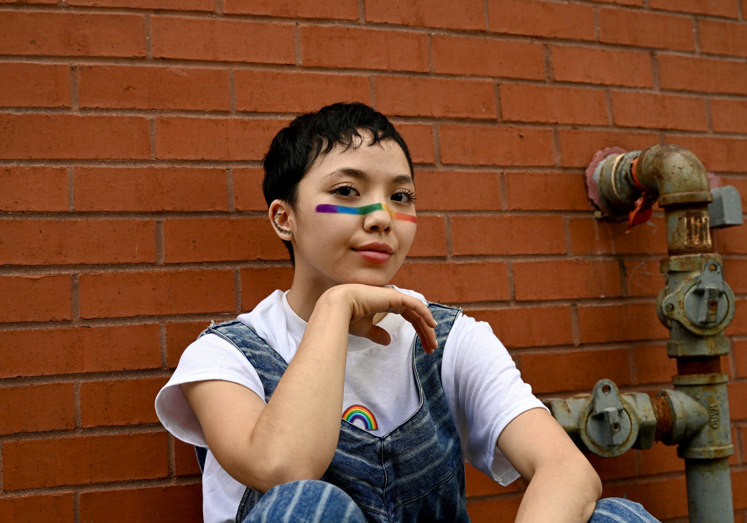 An individual with curly hair and rainbow face paint sitting by a brick wall; exploring gender identity and gender-affirming care.