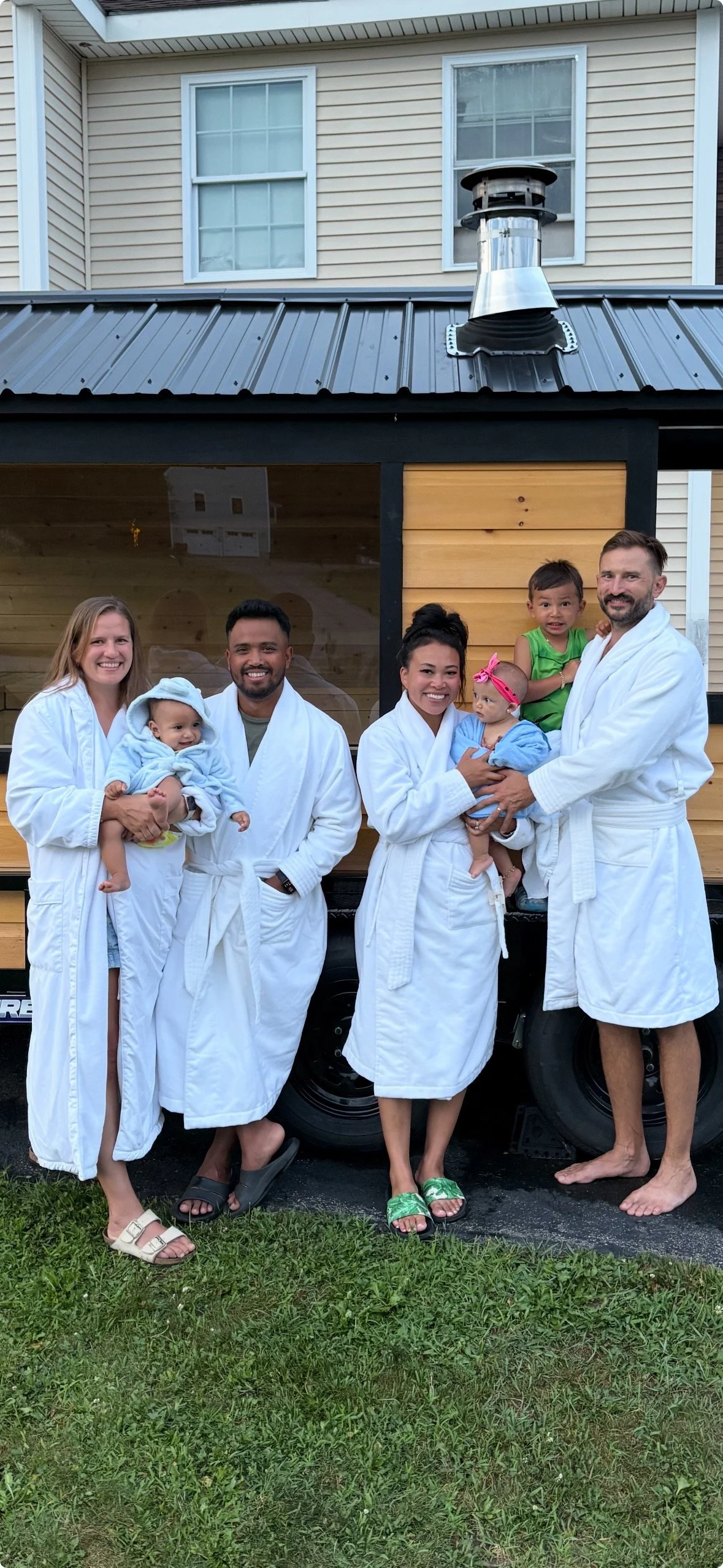 A group of seven people, including adults and children, dressed in white bathrobes, standing outdoors in front of a wooden and glass structure, with a house in the background. They are smiling and some are holding young children.