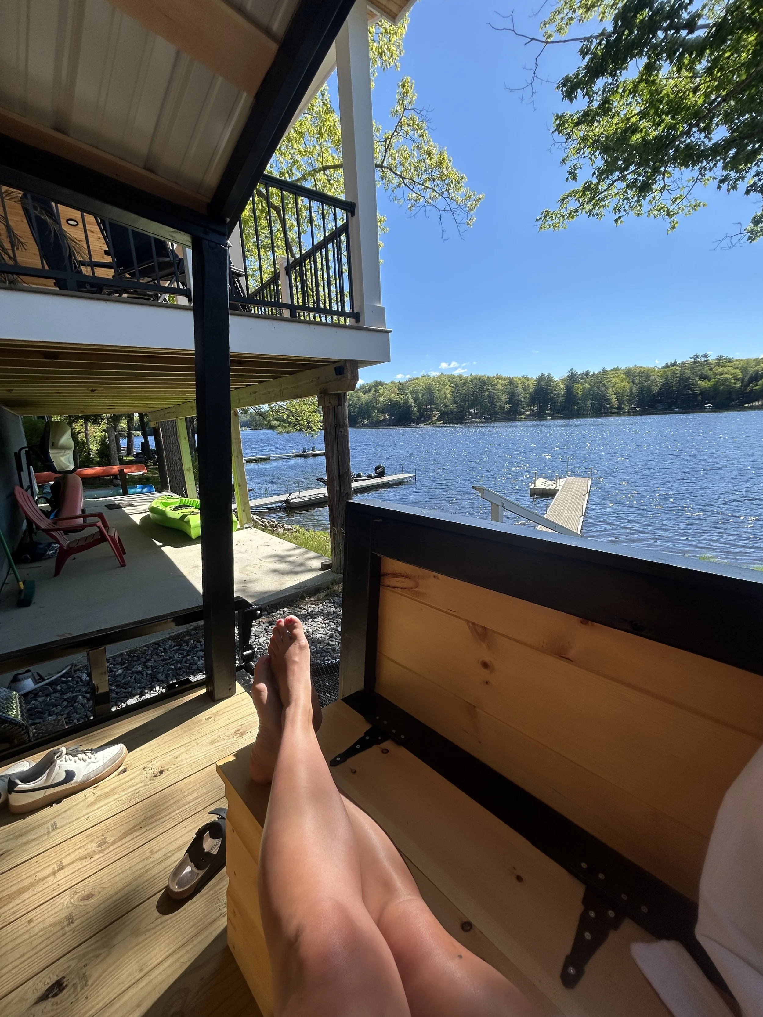 Person relaxing on a porch bench of mobile sauna  by a lake, with their legs stretched out and a view of the water and trees in the background.