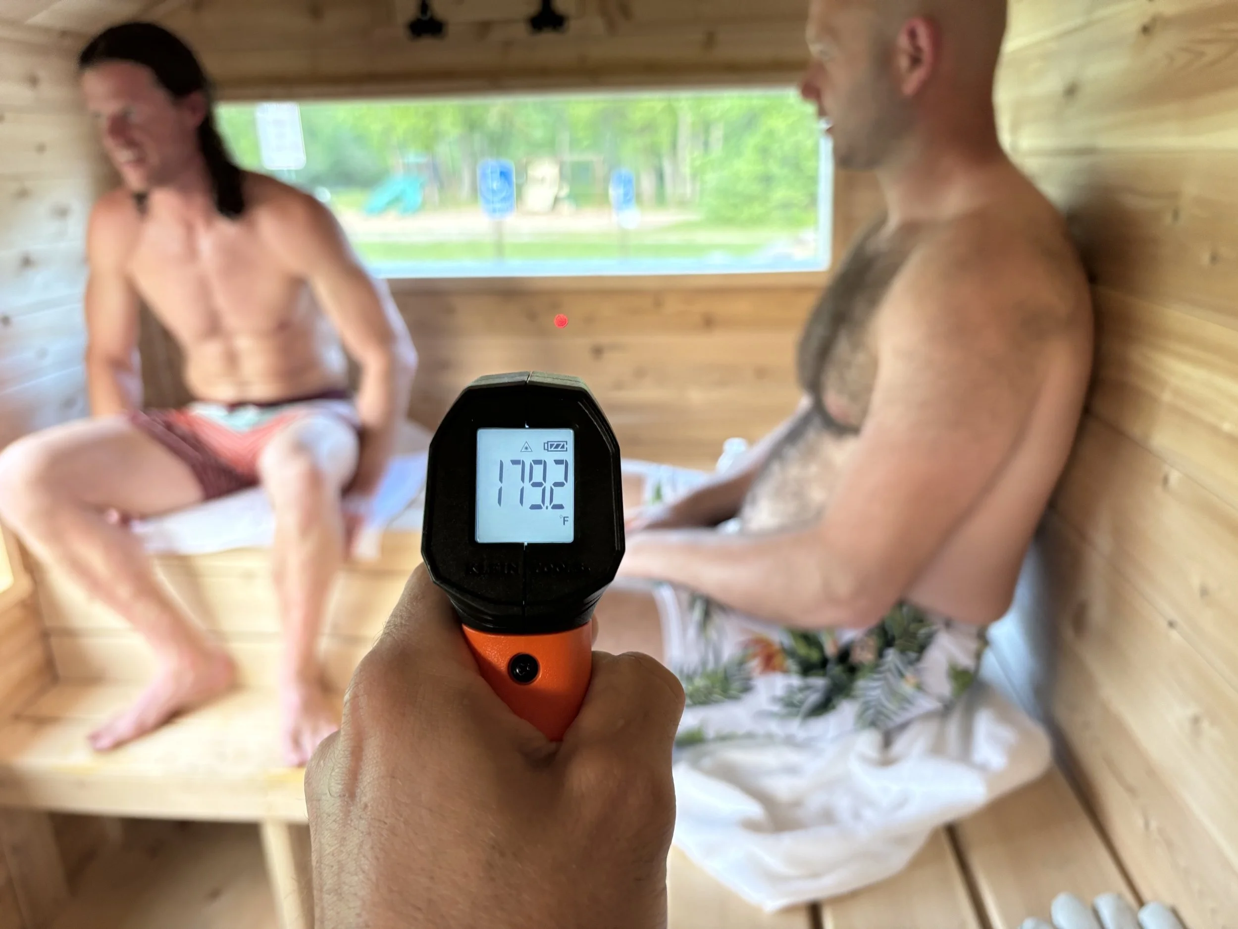 Two men sitting inside a wooden sauna, one with long dark hair and the other bald, both shirtless and wearing swim trunks, while a hand measures their body temperature with a digital thermometer showing 179.2°F.