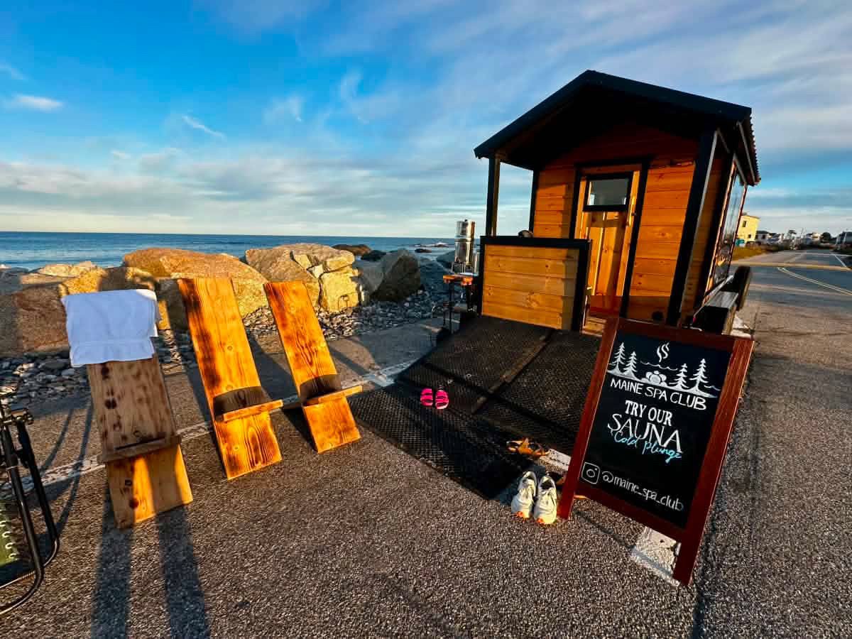 A seaside spa resort with wooden lounge chairs, a small wooden hut, and a sign offering sauna services, with a rocky shoreline and ocean in the background.